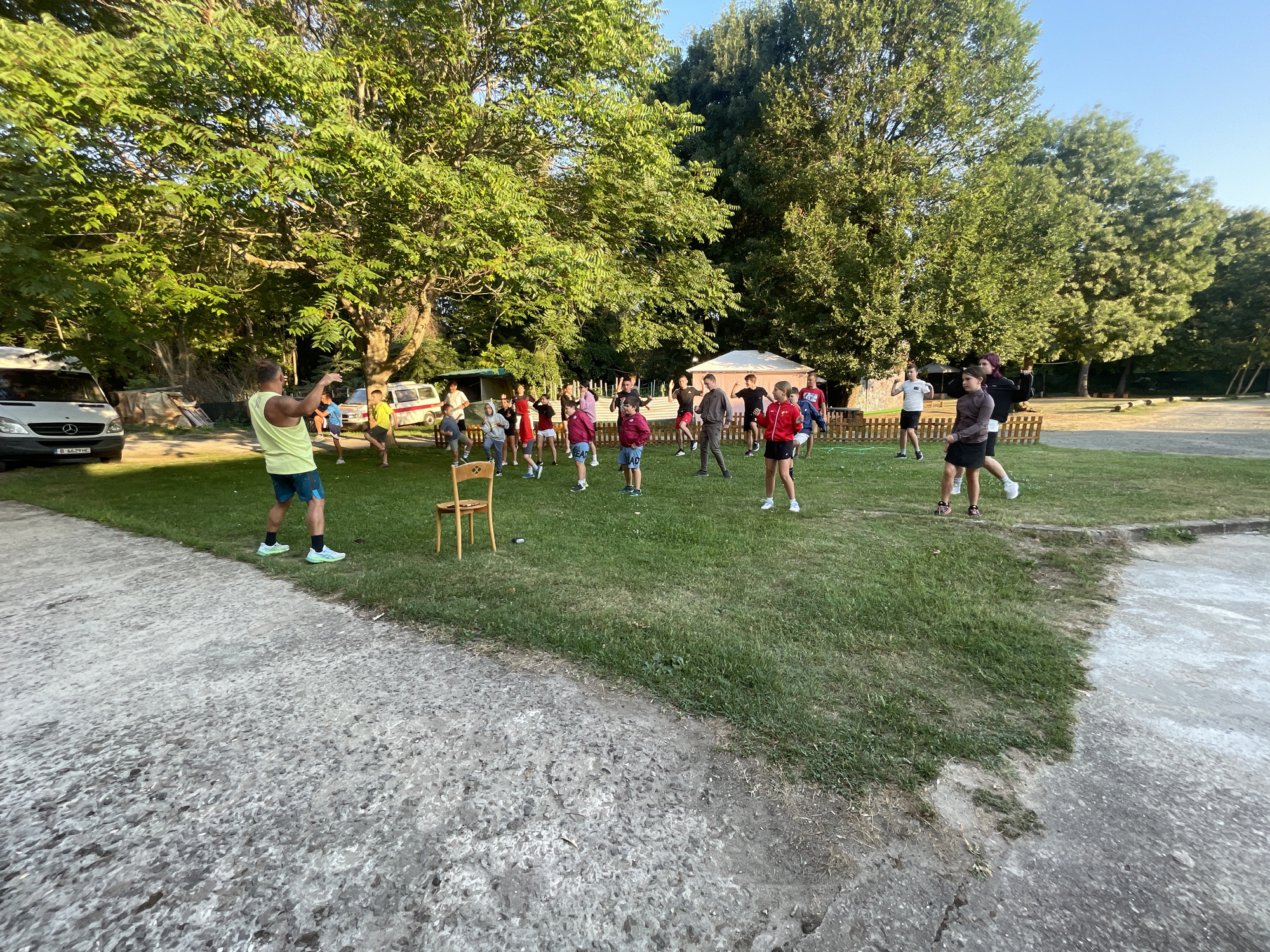 In Primorsko, Bulgaria, a group of approximately 20 people, predominantly young individuals and some adults, are gathered on a grassy area under a canopy of large trees. The scene appears to be outdoors, possibly a campsite or recreational park, with vehicles including a white van visible in the background. The individuals are dressed in casual sportswear and are engaged in what seems to be an organized activity or game, with some holding their hands up in a pose of action. A single wooden chair sits on the grass in the foreground, near a concrete pathway. The lighting suggests it is late afternoon or early evening.
