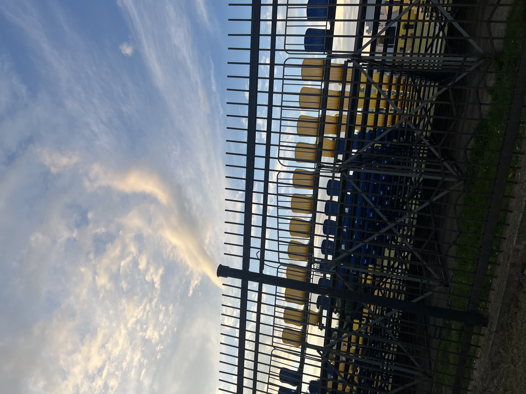 The image shows a view of empty stadium seating under a partially cloudy sky. A black metal fence is in the foreground, obscuring some of the view of the bleachers. The bleachers are a mix of yellow and blue seats. A tall, dark pipe or chimney is visible behind the fence, with a plume of white smoke or cloud extending upwards and to the right. The sky is a bright blue with wispy white clouds, some of which are illuminated by sunlight, giving them a yellowish hue. The image appears to be taken at ground level, looking up at the bleachers and sky. There are no people visible in the image. The setting is likely an outdoor sports stadium or amphitheater. The specific location in the city of Ulyanovsk, Russia is not discernible from the image itself.
