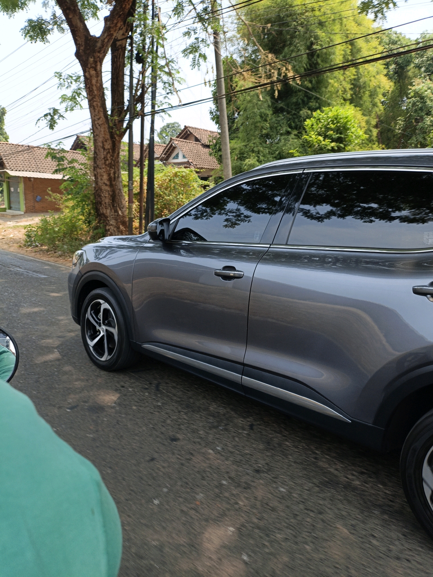 A grey SUV is driving on a road in Cikal, Indonesia. The car is in motion, as indicated by the slight blur in the image. The right side of the SUV is visible, showing its doors, windows, and wheels. The windows are tinted, and reflections of trees and the sky are visible on their surface. The car is driving on an asphalt road, which appears to be in good condition. The surroundings are lush with greenery, featuring trees and bushes. In the background, there are houses with tiled roofs, typical of residential areas in Indonesia. Power lines are also visible, stretching across the sky. In the foreground, a portion of a person wearing a turquoise garment is visible, suggesting that the photo is taken from another moving vehicle, possibly a motorcycle. The scene depicts a common everyday commute or travel scenario in a suburban or semi-rural Indonesian setting.