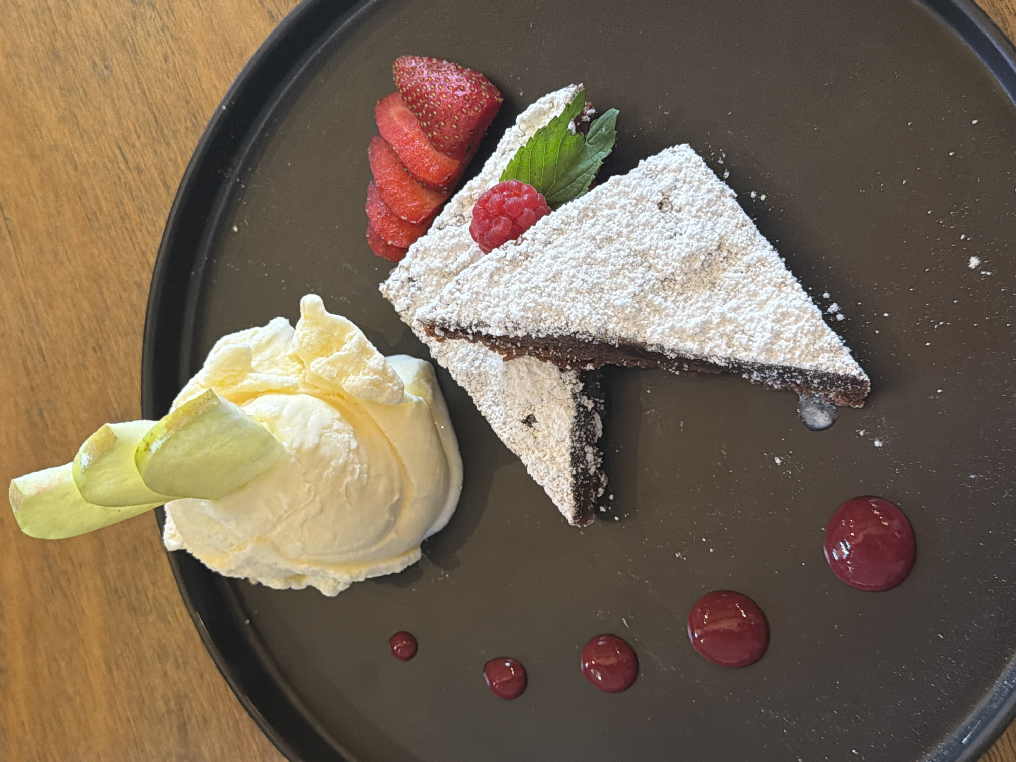 On a black, round plate, a dessert is presented. Two triangular slices of what appears to be chocolate brownie, dusted with powdered sugar, are crossed in the upper-right quadrant. The brownie is garnished with a raspberry, mint sprigs, and a row of sliced strawberries. In the lower-left quadrant, there is a scoop of vanilla ice cream adorned with a curled pale green decoration. From lower-right to lower-left, four circular dots of a dark-red sauce are aligned on the plate. The plate rests on a wood-grained surface. The city of location is unknown.