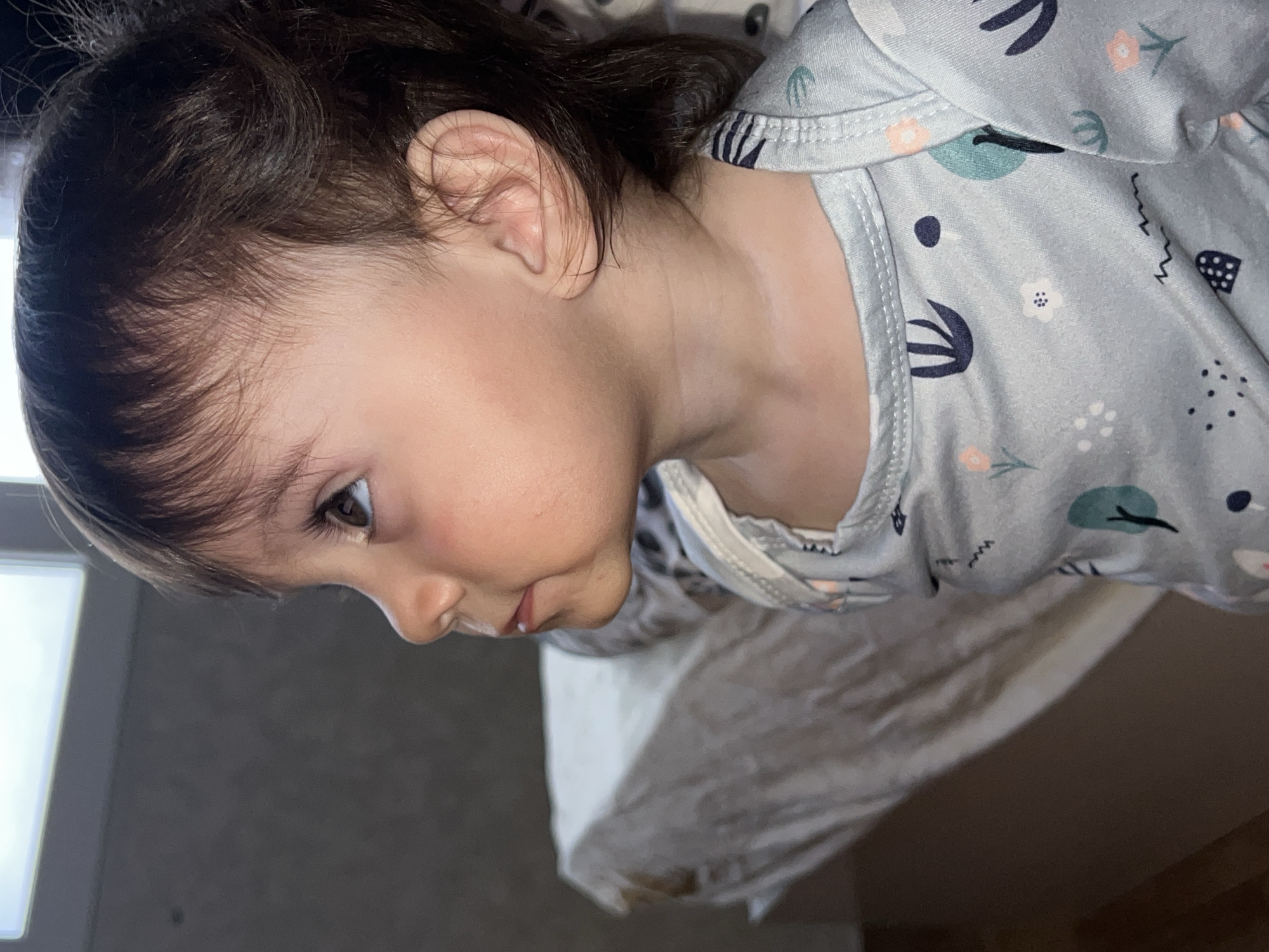 A close-up, slightly tilted shot shows a toddler from the chest up, positioned towards the right side of the frame and gazing downwards. The child has dark brown hair, fair skin, and is wearing a light grey long-sleeved pajama top with a pattern of stylized floral and geometric shapes. The background includes a neutral-colored bedsheet, and a window or light source is visible on the left. There is no indication of the city of Elena, Bulgaria, in the image.