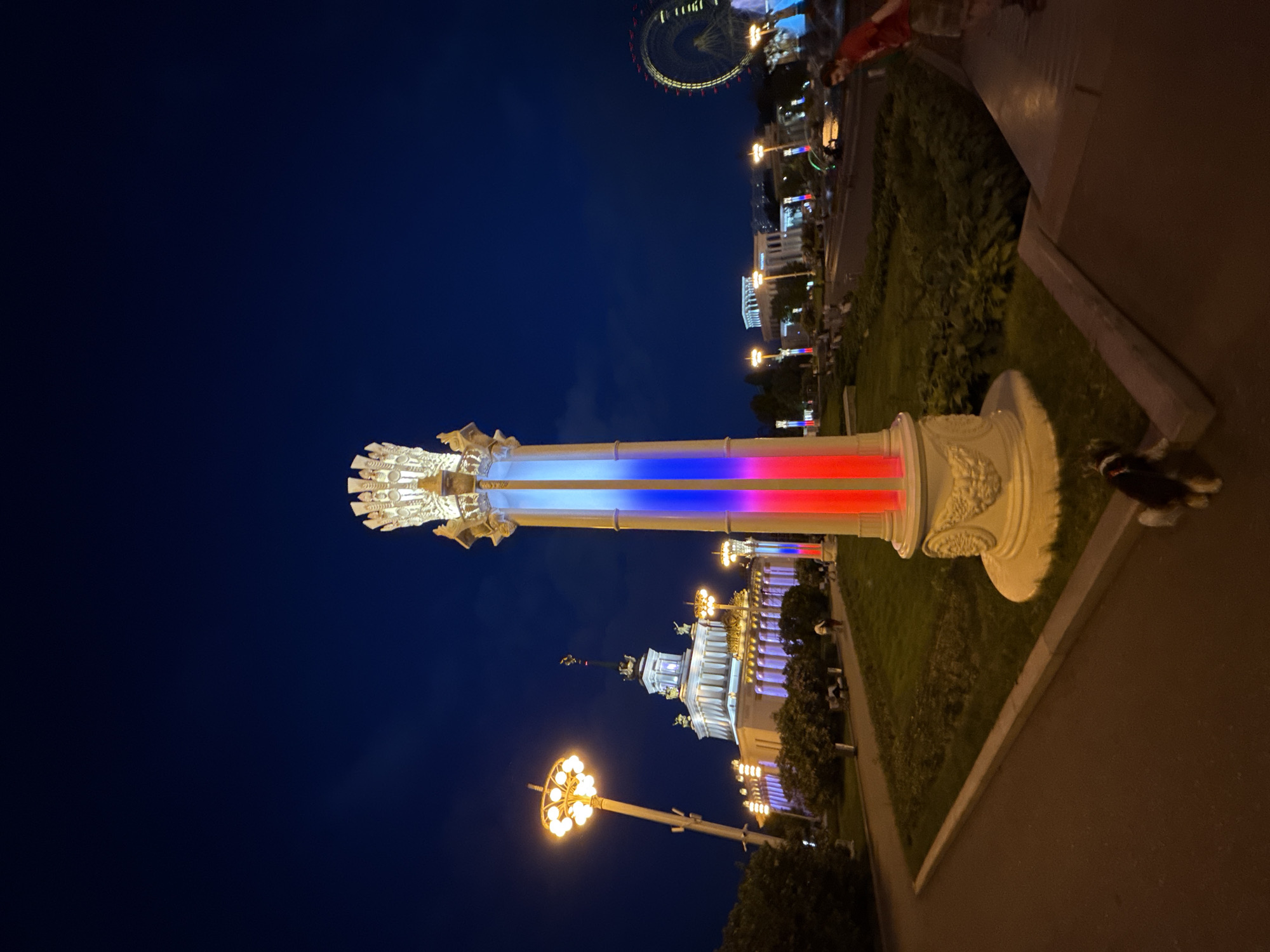 This media file captures a vibrant night scene in Moscow, Russia, likely at a public park or exhibition ground, such as VDNKh. The central focus is a tall, ornate column, brightly illuminated with distinct vertical bands of blue, white, and red light, evoking the colors of the Russian flag. The column features a decorative capital and base, both glowing with warm, golden light.
In the background, a grand, illuminated Ferris wheel with many small lights marks the skyline. Below it, several ornate buildings, possibly pavilions, are visible, also lit up with a mix of warm and cool lights, showcasing their architectural details. Decorative street lamps with multiple glowing spherical lights dot the pathways, casting bright yellow-orange light onto the scene.
The ground consists of paved pathways and manicured grassy areas with bushes and trees. Several people are visible in the distance, strolling along the pathways or near the buildings, enjoying the evening ambiance. In the immediate foreground, a pair of feet wearing white sneakers and dark socks are visible on a paved surface, indicating someone standing close to the camera. The sky is dark blue, characteristic of nighttime, with no visible clouds. The overall atmosphere is festive and lively. There is no visible text in the image.