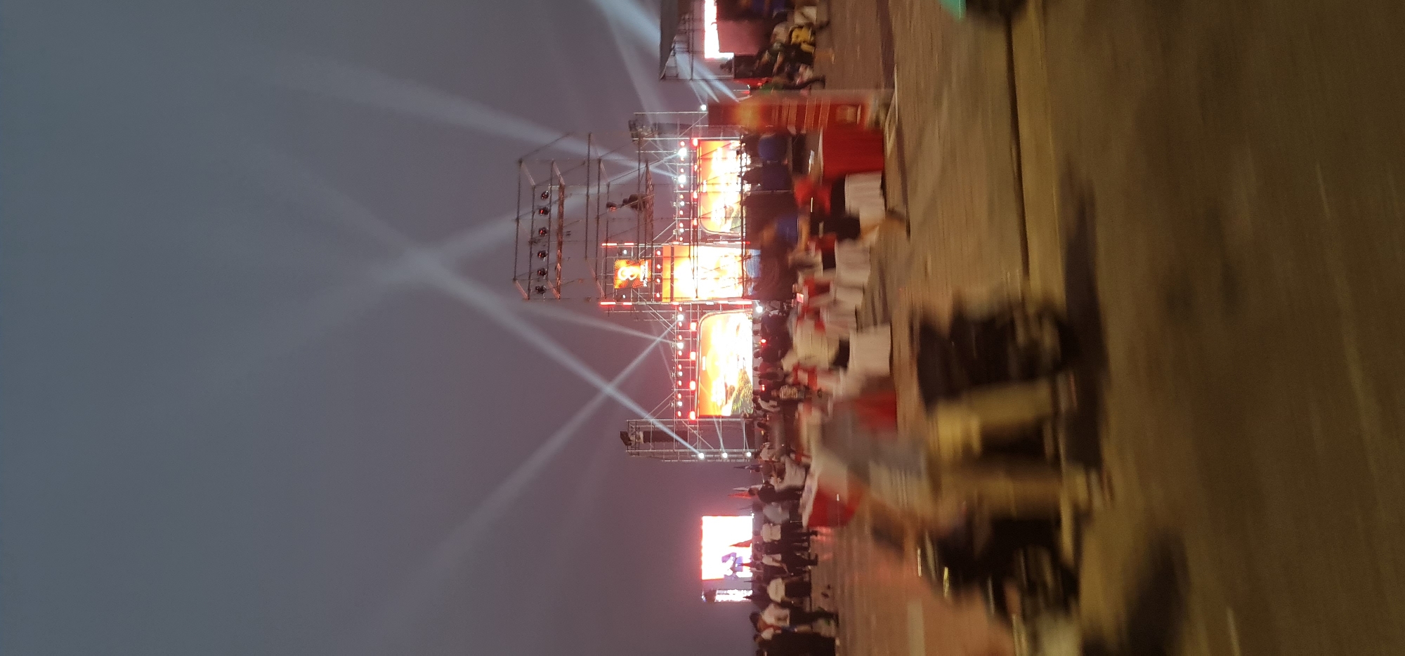 The image depicts a nighttime event, possibly a festival or concert, in Đà Nẵng, Vietnam. A large stage structure is illuminated with bright spotlights, radiating outwards. Several large screens display images. Numerous people are present, some seated at tables with white tablecloths and red accents, possibly VIP seating or dining areas. Many more individuals stand in a crowd closer to the stage. The environment appears to be an outdoor public space or square, with paved ground. Flags are also seen.