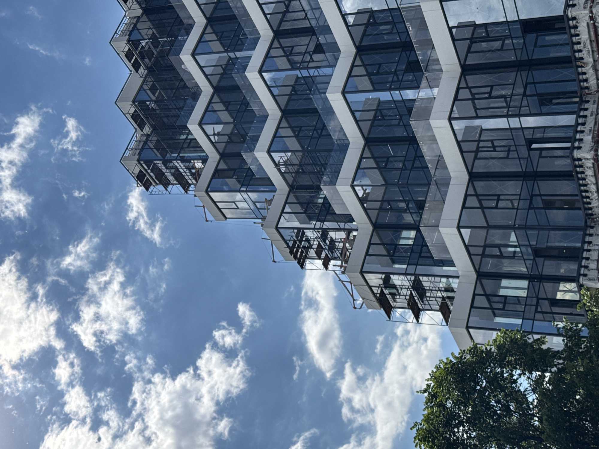 The image, rotated 90 degrees clockwise, showcases a modern building with a distinctive zigzag facade set against a clear blue sky dotted with white, fluffy clouds. The building's complex geometric design features alternating sections of reflective glass and light-colored structural beams, creating a prominent sawtooth pattern that extends dynamically across the frame. Numerous glass panels mirror the vibrant blue sky and scattered clouds, adding depth and visual interest to the structure.
Small elements of scaffolding or construction equipment are visible within some of the building's recessed sections, particularly towards the bottom-left edge of the structure, suggesting ongoing construction or maintenance. A cluster of vibrant green tree foliage occupies the bottom-right corner of the image, providing a natural contrast to the urban architecture. The bright lighting and clear sky indicate a sunny day, likely mid-day or afternoon. Along the far right edge of the building, white material with the repeated text "TENQ" is visible, likely identifying a brand of protective wrap or building material. The scene captures contemporary architecture in Moscow, Russia, under pleasant weather conditions.
