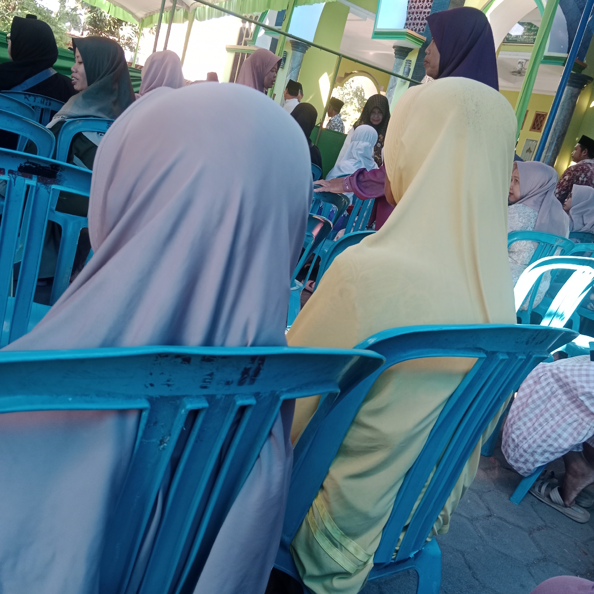 A group of Muslim women, some wearing hijabs, are seated on blue plastic chairs outdoors. The scene appears to be in Tegowanu Kulon, Indonesia, possibly at a community event or religious gathering, indicated by the presence of a green and white tent structure, pillars, and a building in the background that resembles a mosque or community hall. Sunlight illuminates the area, casting shadows on the ground which is paved with stone. Several individuals are partially visible, with their backs to the camera, as well as a few faces in profile and semi-profile. The women are dressed in traditional modest attire. The overall atmosphere seems peaceful and communal.