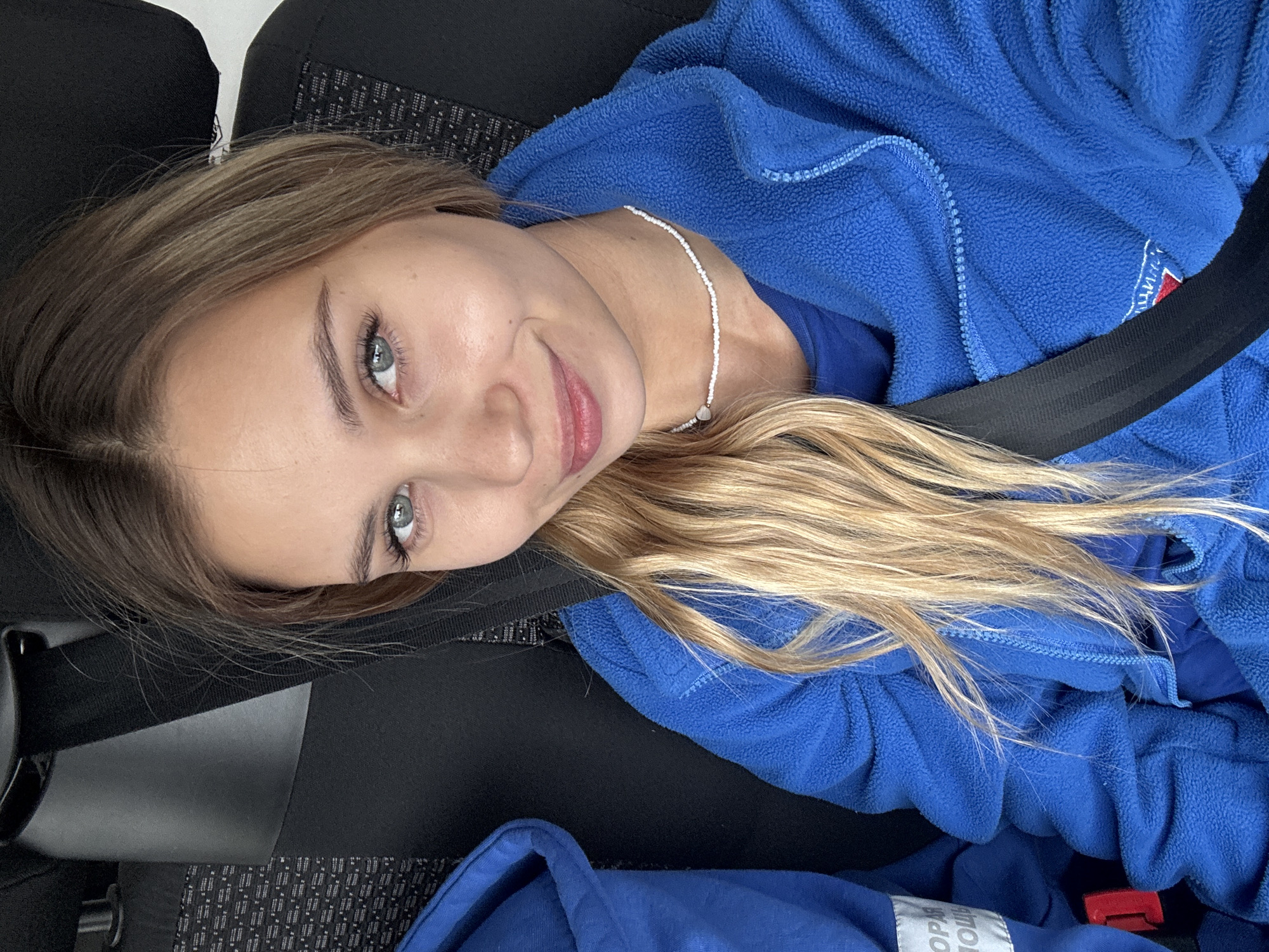 A young woman is seated in a car in Chaikovsky, Russia, wearing a blue fleece jacket, a white beaded necklace, and a seatbelt. She is angled toward the camera, with her blonde hair flowing over the jacket. The car seat has a black fabric pattern.