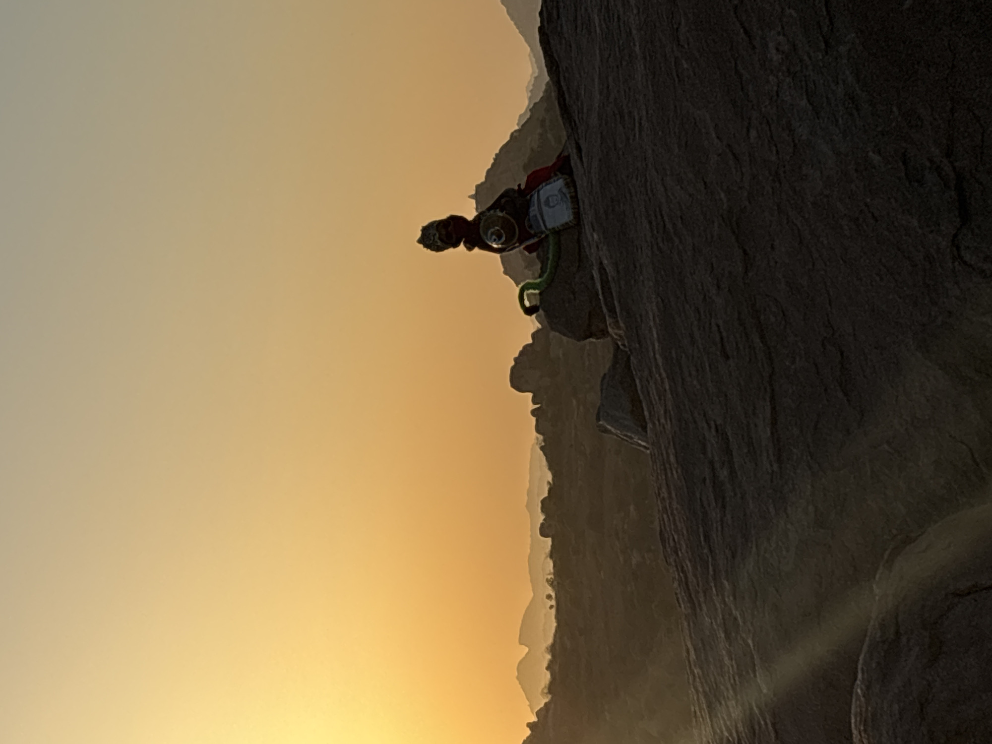 A medium shot shows a rock climber perched on a rock face at Kamalapura, India. The climber, facing the viewer, is wearing a red shirt, and climbing gear, including a helmet, climbing rope, and harness. The climber appears to be preparing for the next move. The rock face is a large, dark stone structure, partially obscured by shadows. The background features a light yellow and orange-hued sky, indicating sunset or sunrise. Rolling hills and other rock formations are faintly visible in the distance.