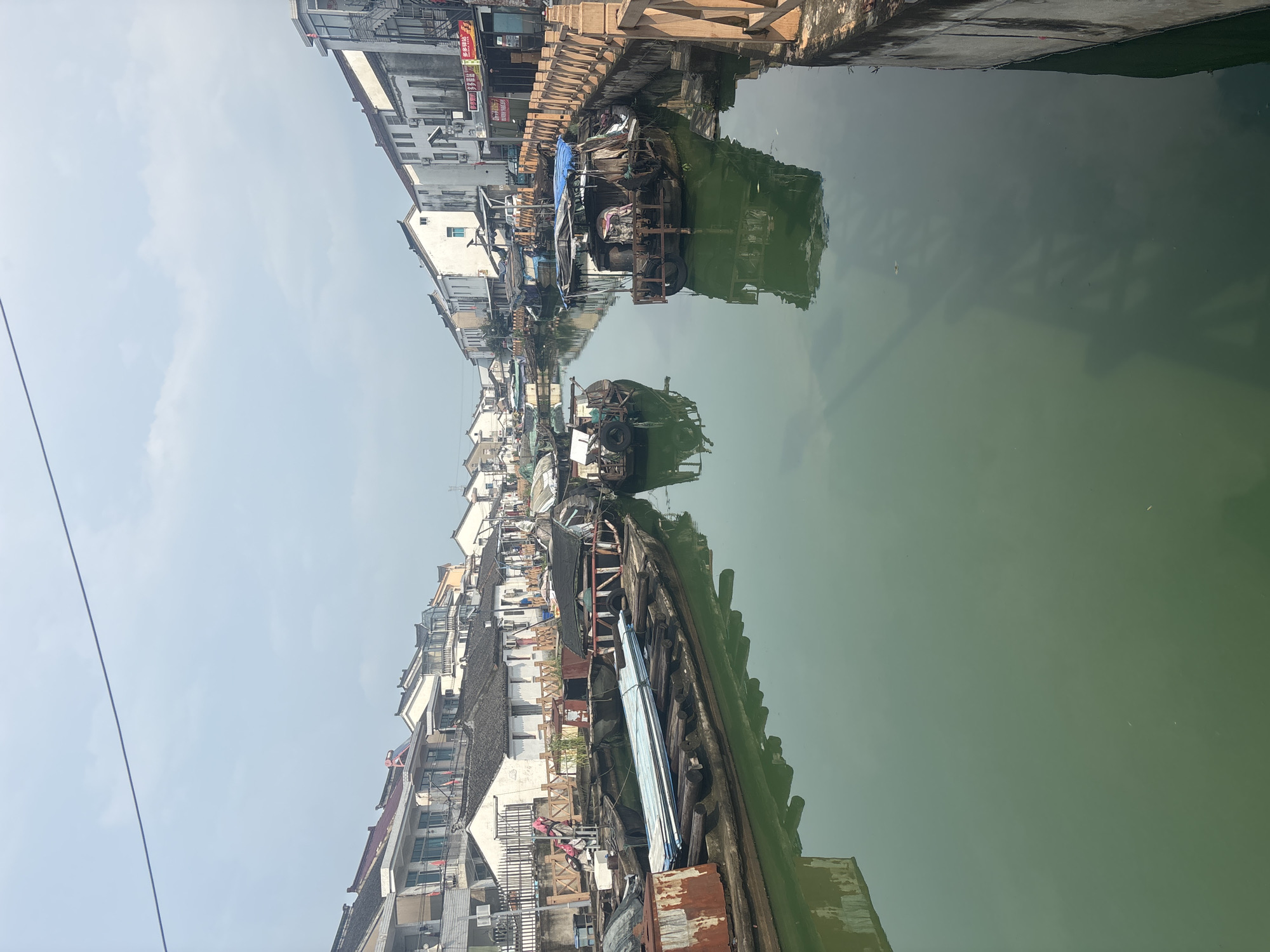 The image depicts a canal scene in Wuzhong District, China, characterized by traditional architecture and a waterway lined with moored boats. The buildings on either side of the canal are predominantly white with dark tiled roofs, some featuring balconies and multiple stories. The canal itself is a murky green, reflecting the overcast sky and the surrounding structures.
Several boats are visible, most of them appearing weathered and used for transport or habitation. Some are laden with what appear to be building materials or salvaged items. The boats are of various sizes and designs, with some covered by tarps.
The canal banks are lined with a wooden railing on the right side of the image, suggesting a pedestrian walkway or viewing area. The water's surface is relatively calm, with reflections of the buildings and boats creating a sense of depth. The overall atmosphere is quiet and somewhat dated, suggesting a historical or less modern part of the district. No people are clearly visible in the image, contributing to a sense of stillness.