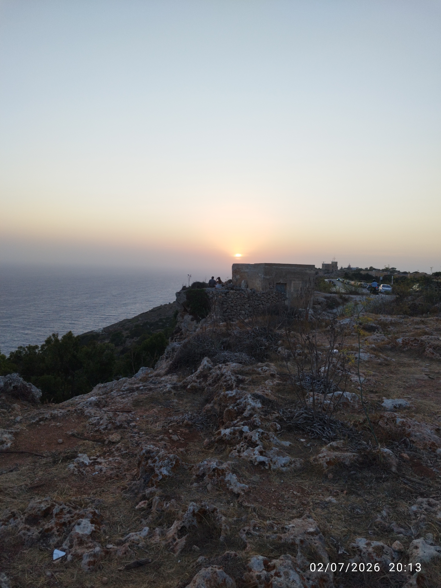 The image depicts a sunset view from Dingli Cliffs, Malta. The sun is low on the horizon, casting a warm glow over the sea. A few people are visible on the clifftop, enjoying the view. In the foreground, the rocky terrain of Dingli Cliffs is evident, with sparse vegetation. In the midground, there is an old stone building, and further in the distance, a road with some cars and another building can be seen. The sky is a pale blue with a hint of orange near the horizon. The timestamp "02/07/2026 20:13" is present in the lower right corner of the image.