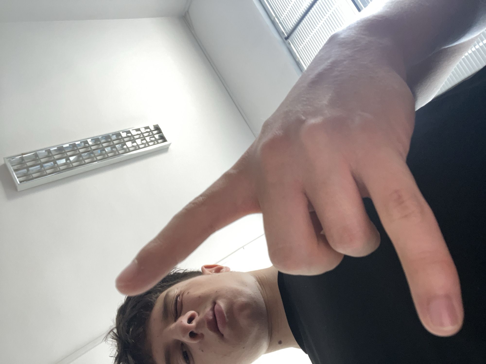 An extreme low-angle, upward-looking selfie depicts a young individual, likely male, with dark, curly hair, positioned in the lower half of the frame. Their face is partially visible, with eyes appearing slightly closed or squinted, and lips subtly parted, conveying a relaxed or casual expression. A large hand, presumably belonging to the subject, dominates the foreground, extended towards the camera with fingers spread, creating a distorted, forced perspective.
The indoor setting in Zlatograd, Bulgaria, features a minimalist design with plain white walls and ceiling. A rectangular fluorescent light fixture is mounted on the wall/ceiling in the upper left background. In the upper right, daylight filters through horizontal window blinds, illuminating the space and indicating it's daytime. The overall environment suggests an office, classroom, or institutional building. No specific activities are depicted beyond posing for the photograph. There is no visible text within the image.