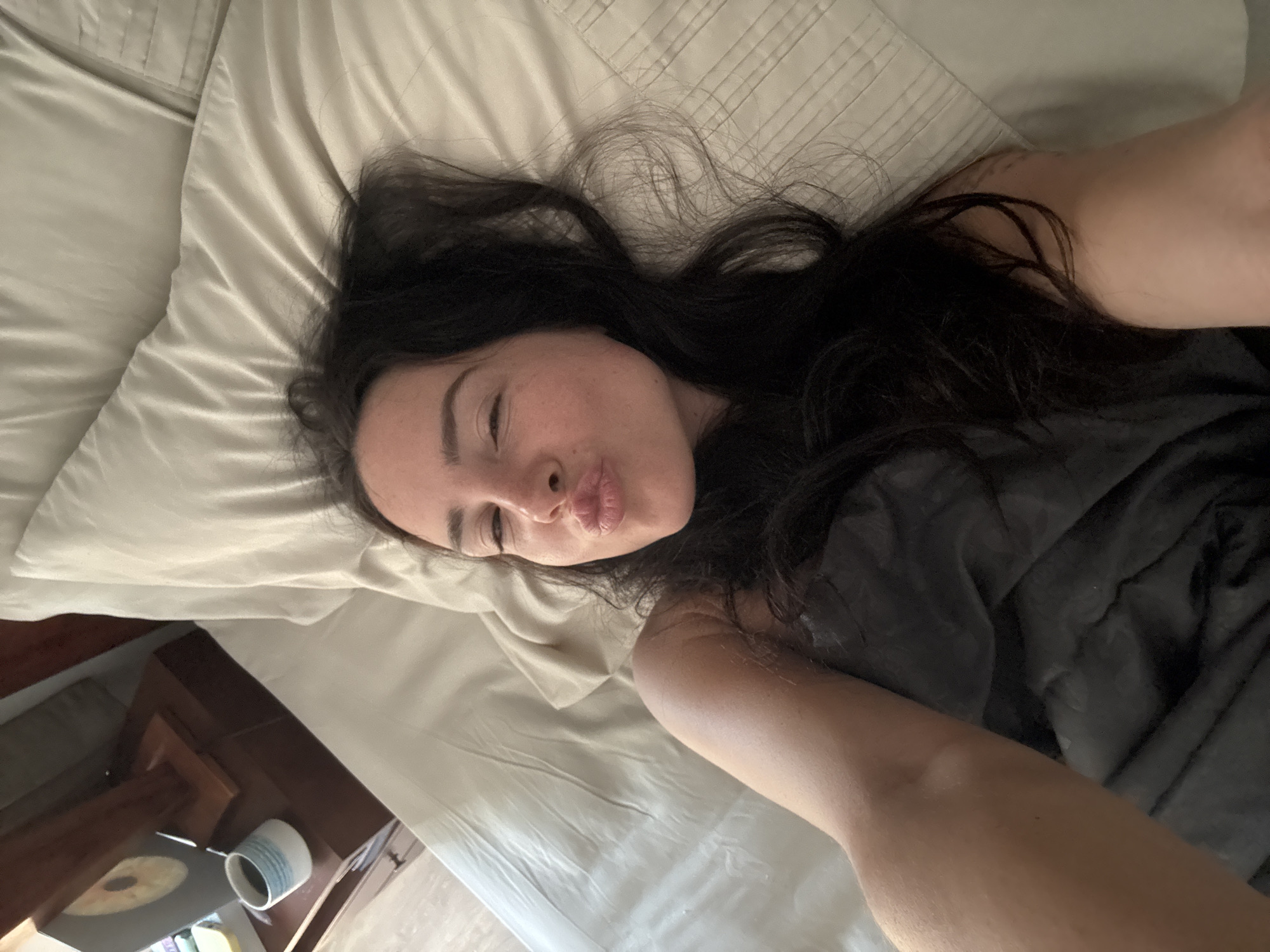 A woman with long, dark hair is lying in bed, taking a selfie. She is puckering her lips and winking one eye. She is covered by a dark gray blanket and is leaning against a light-colored pillow. To the left of the bed, a wooden nightstand is visible with a cup of coffee and a decorative object on top. The environment appears to be indoors, likely a bedroom. This scene could be located in North Kuta, Indonesia, a popular tourist destination known for its beaches and resorts.