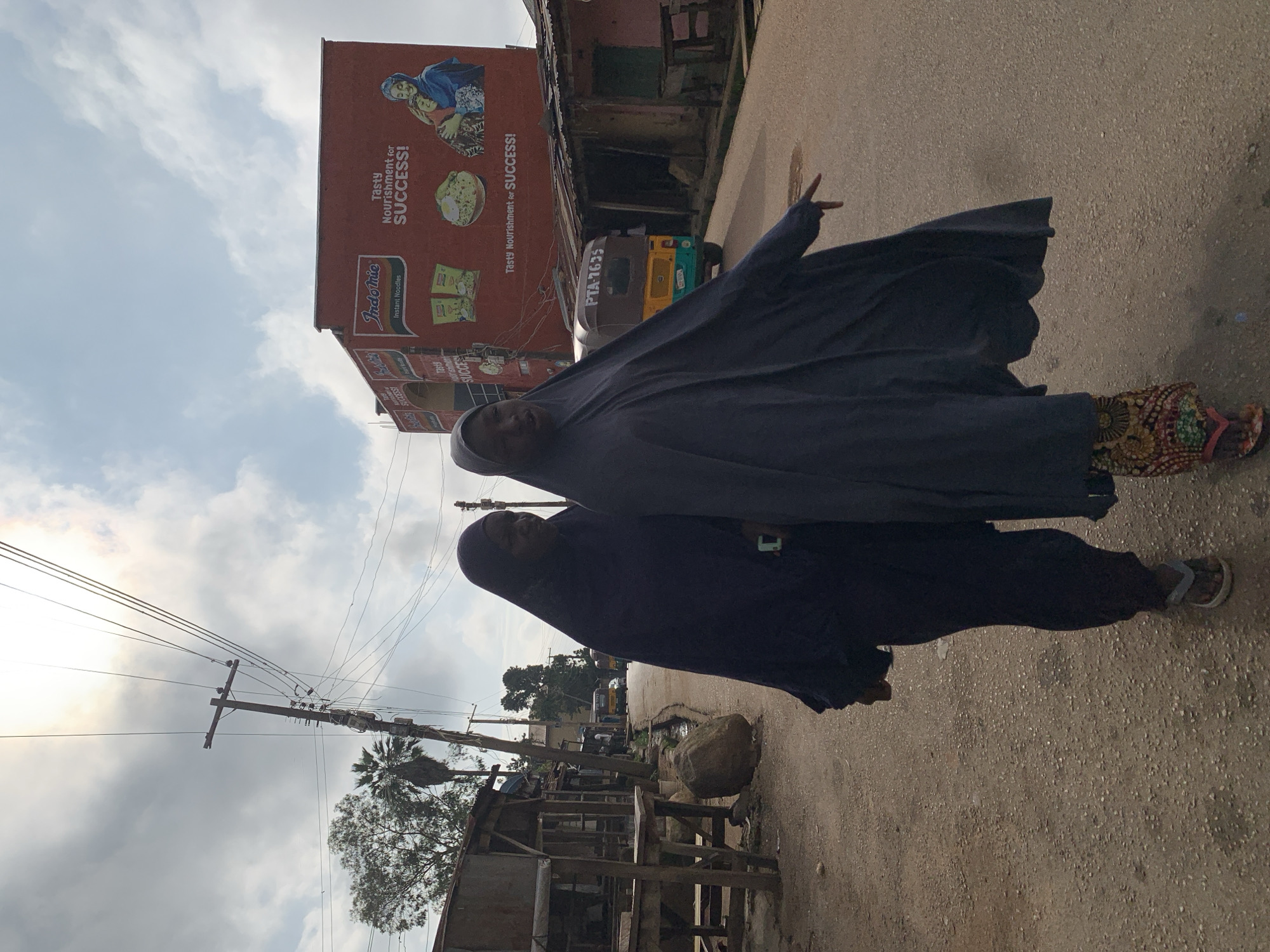 The image depicts two women walking on a dirt road in the city of Jos, Nigeria. They are both wearing hijabs, with one woman in a dark blue hijab and the other in a dark grey hijab. The woman in the grey hijab is pointing with her left hand, and her patterned skirt is visible. Behind them, a bright red building with a large advertisement for Indomie instant noodles is visible. The advertisement features images of the product and a slogan that reads "Tasty Nourishment for SUCCESS!". A yellow and green auto-rickshaw with the license plate "PTA 7635" is also present on the road, along with a utility pole with numerous wires. The environment appears to be a street with modest buildings and a somewhat unpaved road, suggesting a local urban setting. The sky is cloudy with the sun partially visible.