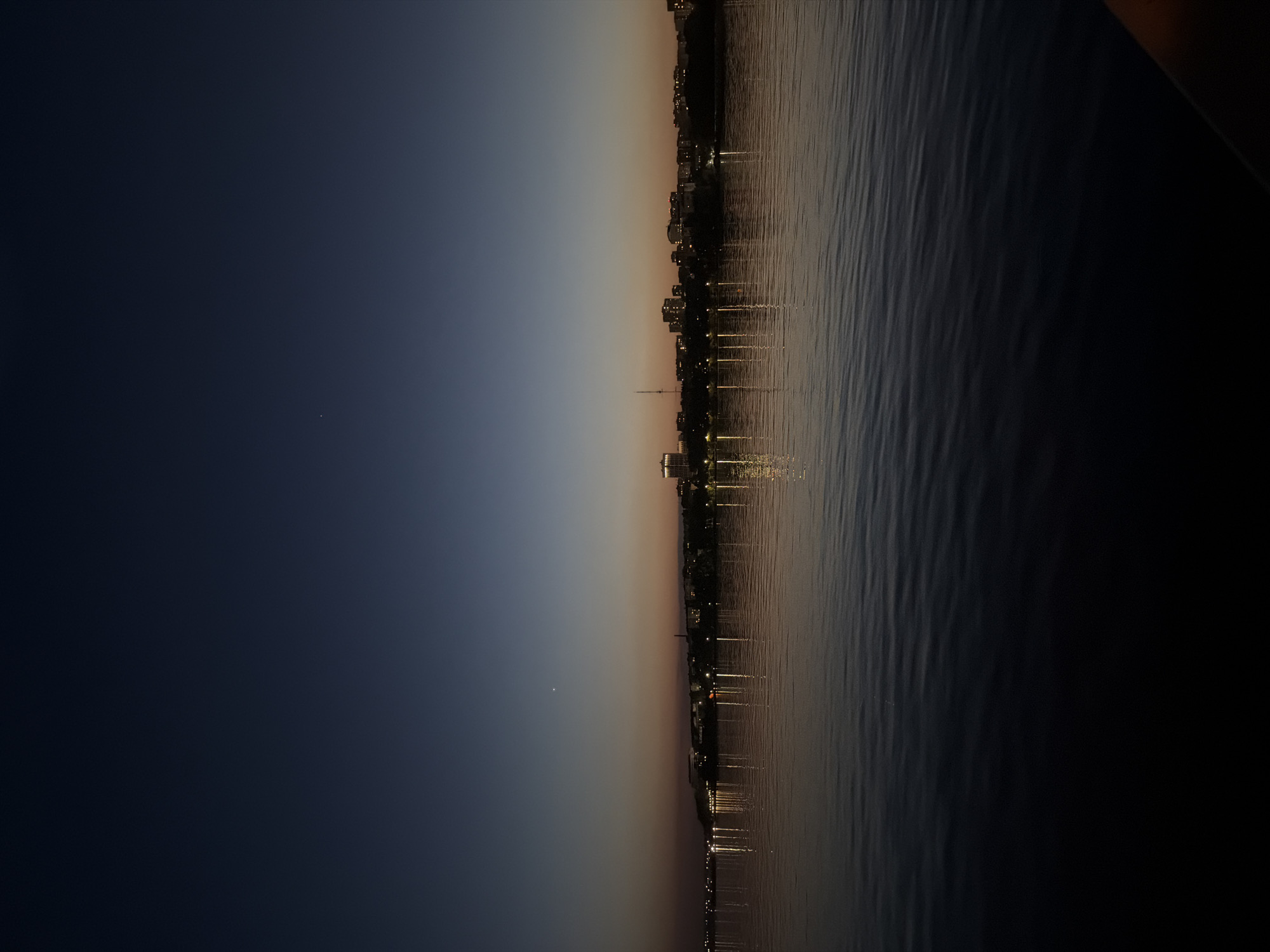 The image depicts a nighttime view of a city skyline reflected in a body of water, likely a lake or river. The sky transitions from dark blue on the left to a gradient of orange and pink near the horizon. A small, bright spot, possibly a star or planet, is visible in the dark blue sky. The city skyline consists of buildings and structures silhouetted against the sunset colors. Lights from the buildings are reflected as vertical streaks in the water, creating a mirrored effect. The water surface shows ripples or small waves. The specific location is Izhevsk, Russia.