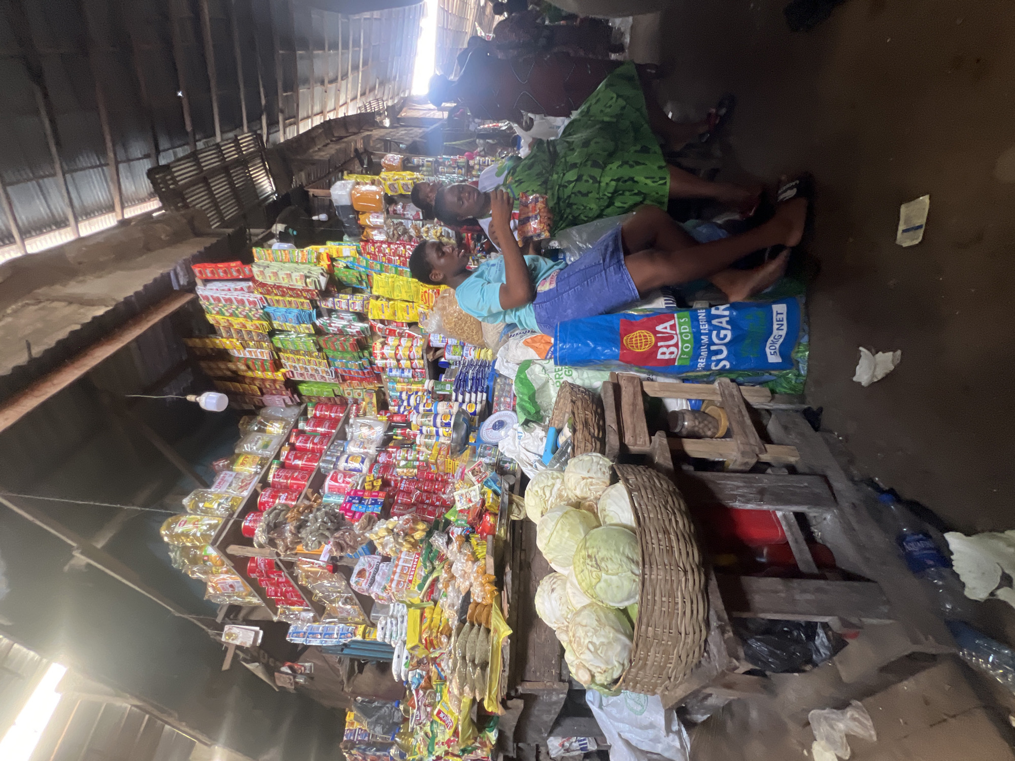 The image depicts a market scene in Nkpor, Nigeria. Two individuals are lying on sacks labeled "BUA Foods Premium Kernel Sugar 50kg Net," seemingly taking a rest amidst the market activity. A third person is visible nearby. Stacks of various goods, including packaged foods and household items, are arranged behind them, suggesting a vendor's stall. A basket overflowing with cabbages is positioned prominently in the foreground. The market area is under a corrugated metal roof, and the environment appears somewhat cluttered and dimly lit.