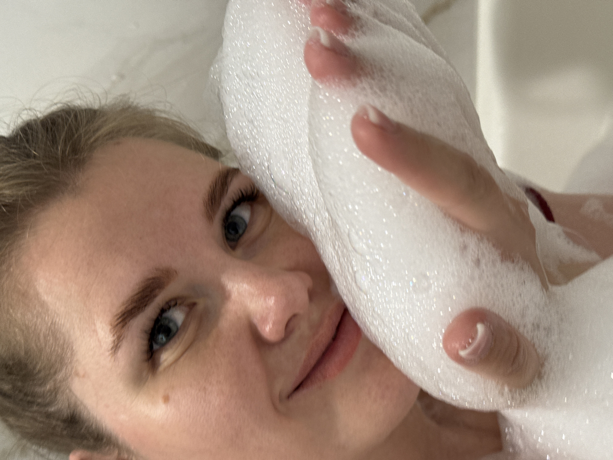 A young woman with light skin and blue eyes is lying down, holding a mass of white soap foam near her face. The image is likely taken indoors, possibly in a bathtub or shower. The location is Ферма, Russia.
