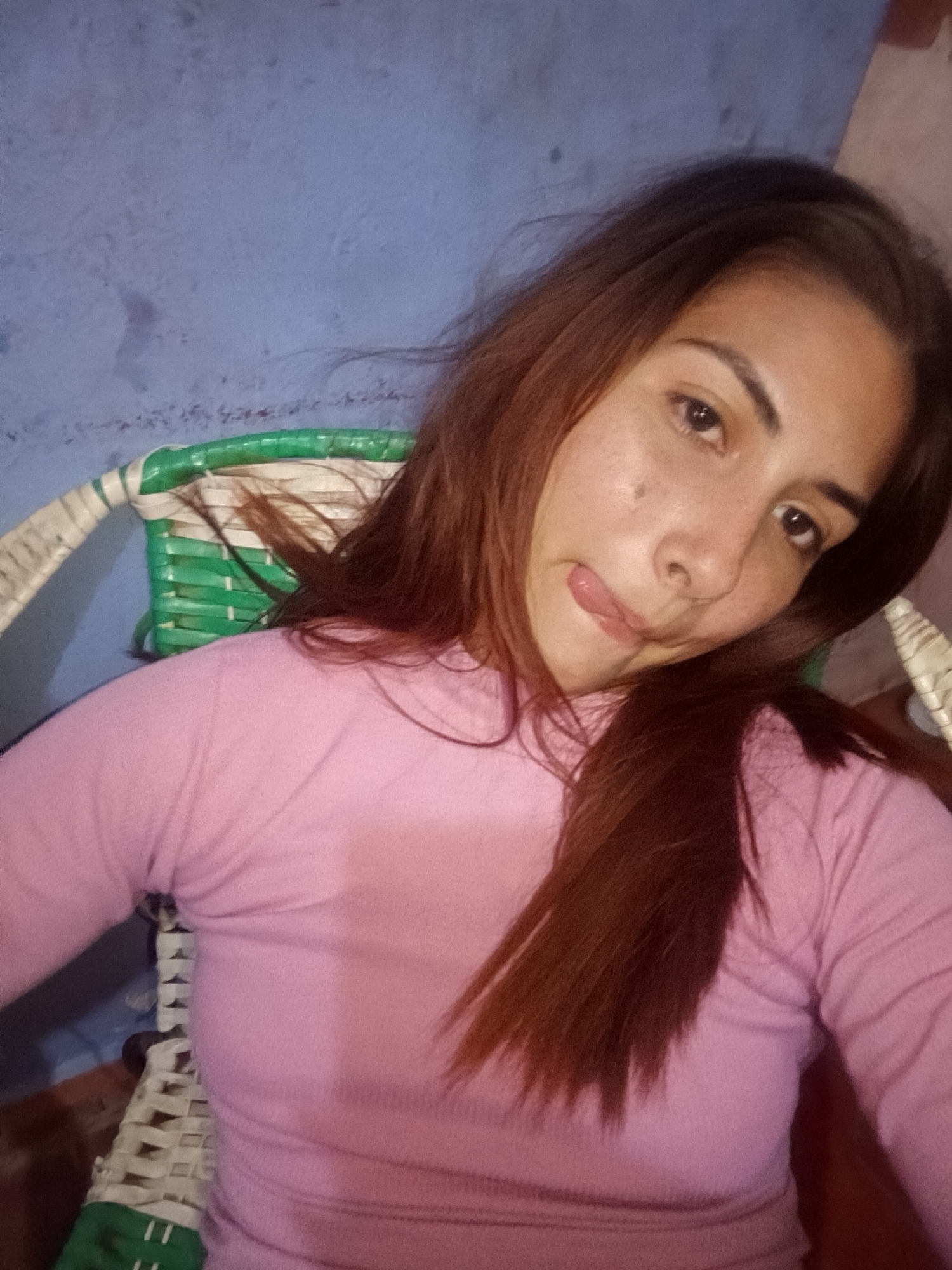 A light-skinned woman with long, reddish-brown hair is seated in a white and green woven chair against a blue wall. She is wearing a pink long-sleeved shirt and is sticking her tongue out.
The location is Juan Griego, Venezuela.