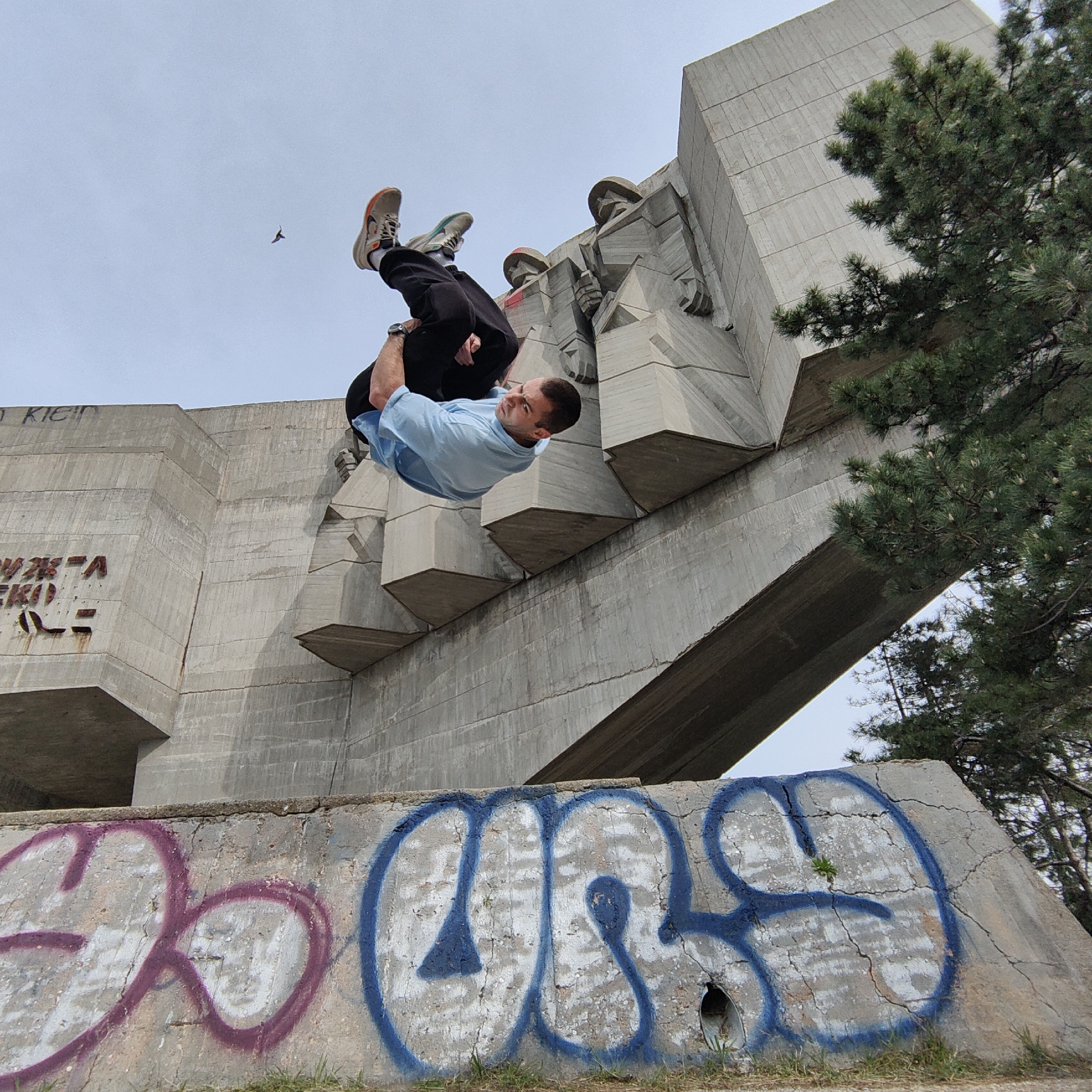 The image depicts a man performing a backflip in front of the Monument to the Soviet Army in Varna, Bulgaria. The monument is a large, brutalist concrete structure featuring sculpted figures. Graffiti is visible on the lower portion of the monument. The man is wearing a light blue shirt and dark pants. His legs are bent, and his arms are tucked in. A bird is flying in the sky. A pine tree is in the foreground. The sky is overcast.