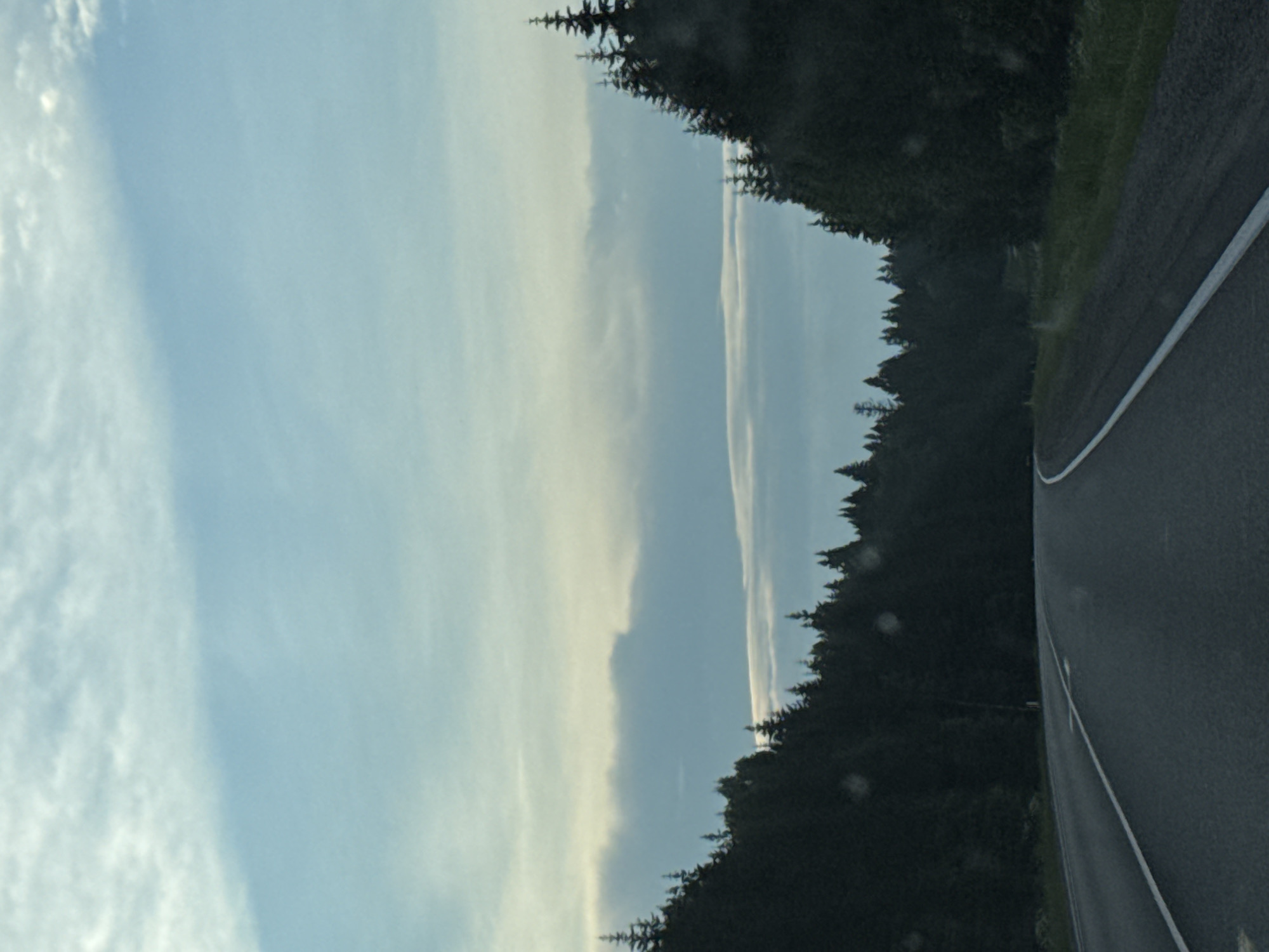 The image shows a winding road through a forest. The sky is overcast with scattered clouds. The trees are dark green and appear to be coniferous. The road is paved and has a white line marking the lane. The overall impression is one of a rural or wilderness setting. The image is a photograph taken from a moving vehicle, as indicated by the slight blur and the perspective. There are no people or animals visible in the image.