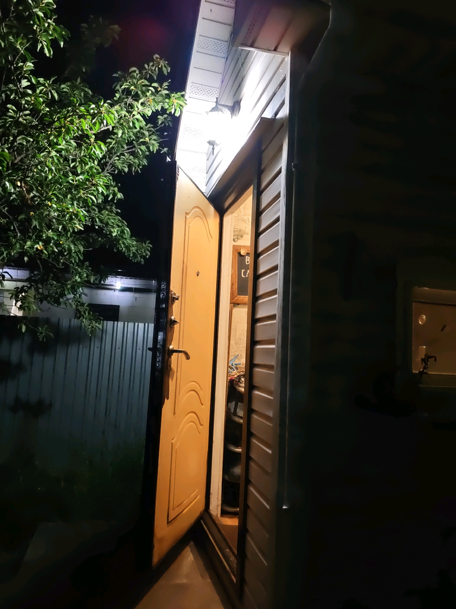 The image depicts an exterior view of a partially opened yellow door at night. The door has a textured surface with curved designs and features two handles and a keyhole. Beyond the open door, a dimly lit interior is visible, with shelves and objects suggesting a storage area or entryway. The exterior wall of the building is covered in horizontal siding. To the left of the door, a large green tree with dense foliage is prominent, its leaves illuminated by an unseen light source. In the background, a grey metal fence runs horizontally. A light fixture is mounted above the door. The scene appears to be a residential or utility area, possibly in a backyard or alley. The image is shot at a low angle, looking upwards towards the door and the tree.
