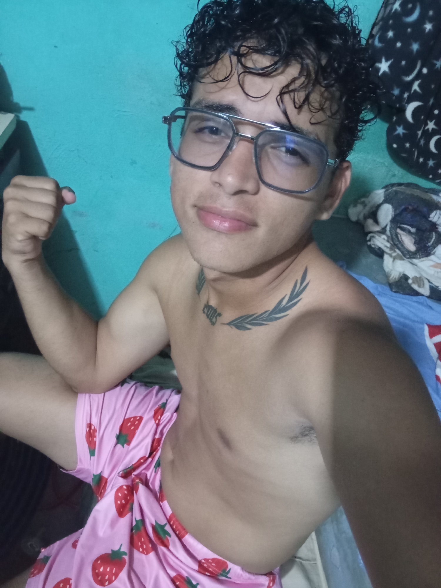 A young individual, appearing male, is seated indoors, looking directly at the camera with a slight smile. He has dark, curly hair and wears silver-framed, slightly rectangular glasses. He is shirtless, revealing his bare torso, and is wearing pink shorts with a repeating red strawberry pattern. His left arm is bent at the elbow with the hand clenched into a fist. Two tattoos are visible: one on the left side of his neck and upper chest, resembling a stylized dark leaf or laurel branch, and another dark tattoo below it on the clavicle area, which appears to be text or numbers. Armpit hair is visible under his right arm. The background on the left consists of a solid teal wall, with a light-colored object and a dark cable. On the right, there is dark fabric patterned with white stars and crescent moons, a blue sheet, and some crumpled fabric, indicating a domestic setting like a bedroom.