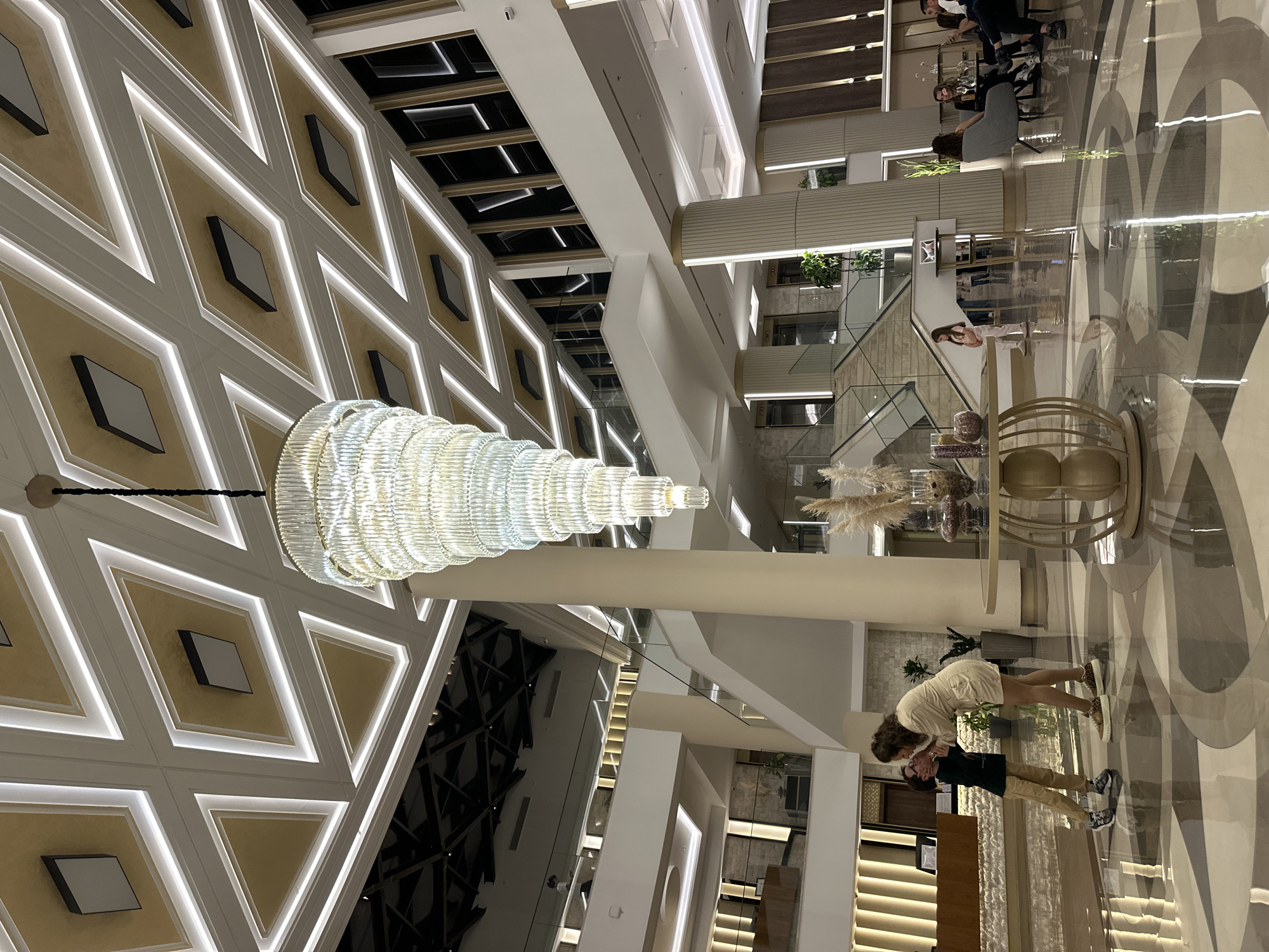 This media file captures the grand interior of a modern, multi-level building, likely a hotel lobby or atrium, in Kazanlak, Bulgaria. The perspective is slightly elevated, looking across the expansive space.
The ceiling features a striking geometric design of alternating light-colored diamonds and triangles, outlined by white borders with integrated strip lighting, set against a subtly textured, warm-toned background. A magnificent, multi-tiered crystal chandelier, composed of numerous stacked glass rings, hangs prominently in the upper-left area.
The floor is highly polished and reflective, mirroring the architectural elements and people. It exhibits a large, intricate pattern of sweeping curves and geometric shapes in shades of gray, white, and beige. The building's structure includes multiple levels connected by sleek staircases, some with glass railings, and elevated walkways, all bathed in soft artificial lighting.
In the lower-right foreground, a couple is captured in a tender moment. A woman with dark, curly hair, wearing a light-colored top and shorts with leopard-print shoes, leans down to kiss or hug a man dressed in a dark t-shirt, light pants, and sneakers. Their clear reflections are visible on the glossy floor beneath them.
In the mid-ground and background, several other people are visible on various levels. Some individuals are seated at modern lounge tables, appearing to socialize, while others are standing or moving through the space. A large decorative table with a gold-toned base, adorned with pampas grass and other ornaments, stands nearby.
The scene is illuminated primarily by artificial light sources from the ceiling patterns, the chandelier, and integrated architectural lighting, suggesting an evening or nighttime setting. The atmosphere appears sophisticated and serene. There is no visible text in the image.
