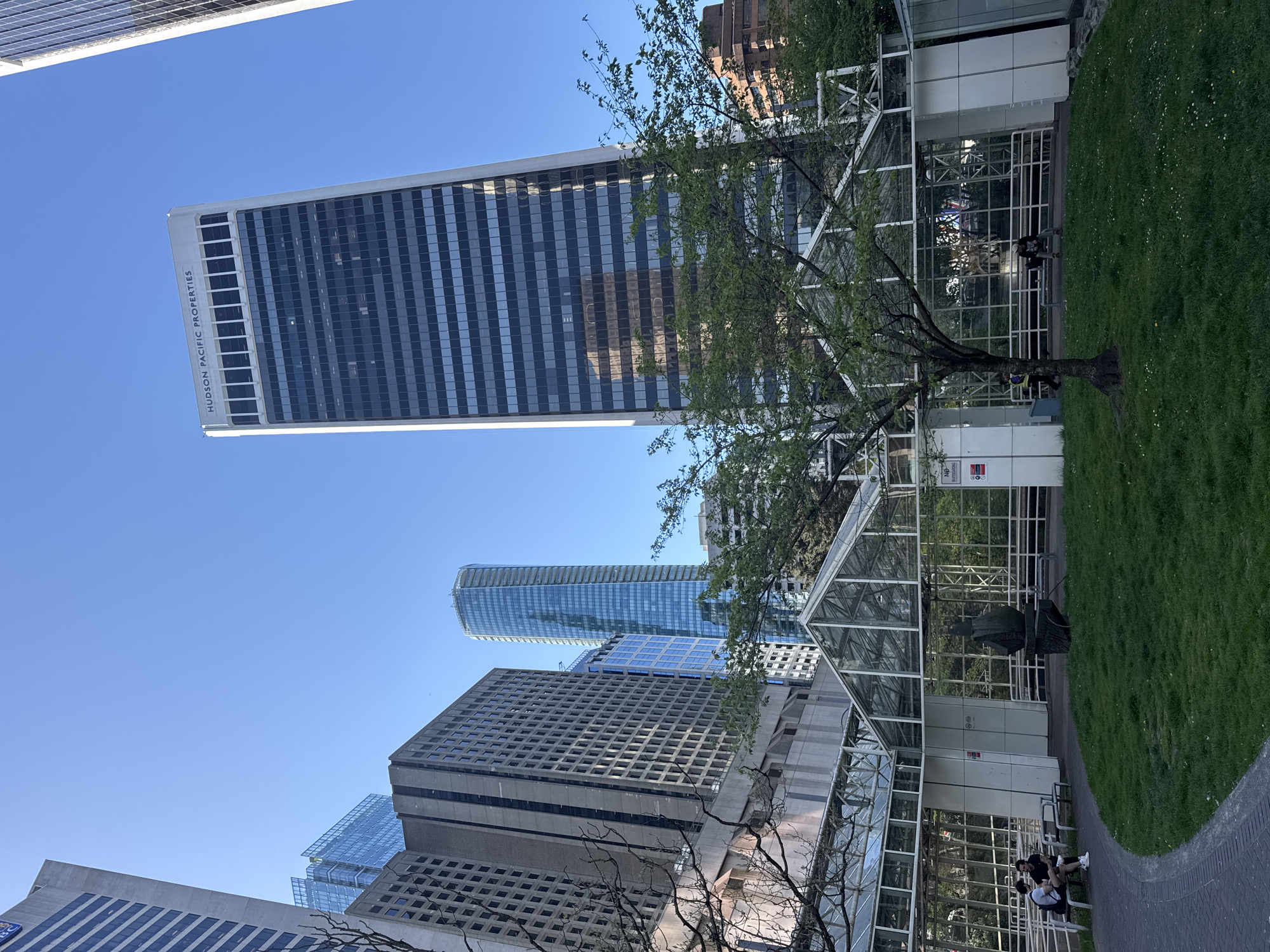 This media file presents a vibrant daytime scene in Vancouver, Canada, showcasing a blend of modern urban architecture and green public space under a clear blue sky.
A prominent feature is a tall, modern skyscraper with a reflective glass facade and a distinctive grid pattern, located on the left side of the frame (when rotated to upright orientation). Vertically displayed on its side is the text "HUDSON PACIFIC PROPERTIES." Several other high-rise buildings populate the background and midground, including a sleek, curved skyscraper with blue glass and other concrete and glass structures, contributing to the city's dynamic skyline.
In the foreground and right side of the frame, a lush green park area unfolds, featuring a paved pathway. Within this park is a large glass structure, likely a conservatory or greenhouse, characterized by a white grid framework. Trees with both verdant foliage and more skeletal branches are scattered around the park.
A few people are visible enjoying the park. Most are seated on benches along the paved path next to the glass structure, appearing relaxed. A dark, abstract sculpture or statue is discernible inside the glass building. Near the glass structure, a white sign bears some unreadable text, though a red "NO SMOKING" symbol is partially visible. The clear blue sky and bright light suggest a sunny day, creating a peaceful and pleasant atmosphere.