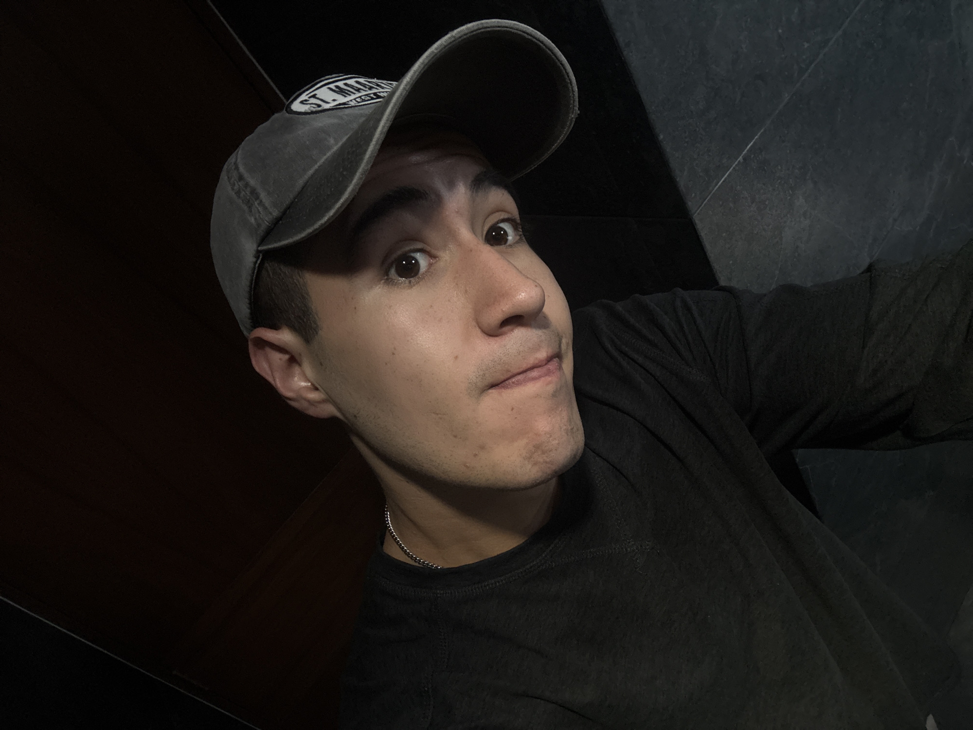 The image is a close-up selfie of a young man, likely taken indoors in San Bernardino Tlaxcalancingo, Mexico. He looks directly at the camera with wide, dark eyes and a slightly surprised or curious expression, his lips forming a subtle smirk or pout. He wears a faded dark grey baseball cap with a white oval patch on the front; visible text on the patch includes "ST. MAA" and "WEST". His short dark hair is visible beneath the cap.
The man is dressed in a dark, possibly black, crew-neck t-shirt, and a thin silver chain necklace is visible around his neck. The lighting is dim, suggesting an evening or nighttime setting. The background on the right is a dark grey tiled or paneled wall with faint lines, while the left side is an even darker, indistinct surface, possibly a shadowed wall or piece of furniture. The overall impression is a casual, direct portrait with an engaging, somewhat playful expression.