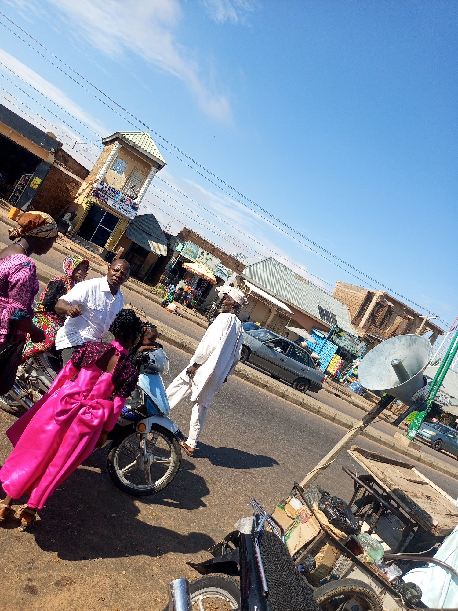 The image shows a street scene in an urban environment. In the foreground, a group of people are gathered around a motorcycle. A woman in a bright pink dress is leaning against the motorcycle, and a man in a white shirt is standing next to her, appearing to be talking to her. A young child in a light blue outfit is also present near the motorcycle. Behind them, several other people are visible, some walking and others standing.
The background consists of buildings lining the street, suggesting a commercial area with shops. There are also vehicles on the road, including cars and another motorcycle. A large, metallic loudspeaker is mounted on a pole, and a cart filled with various items, including boxes and trash bags, is also visible. The sky is clear and blue, with a few scattered clouds. The overall impression is a busy street scene, possibly in a market area. The specific location is identified as Unknown, Unknown.