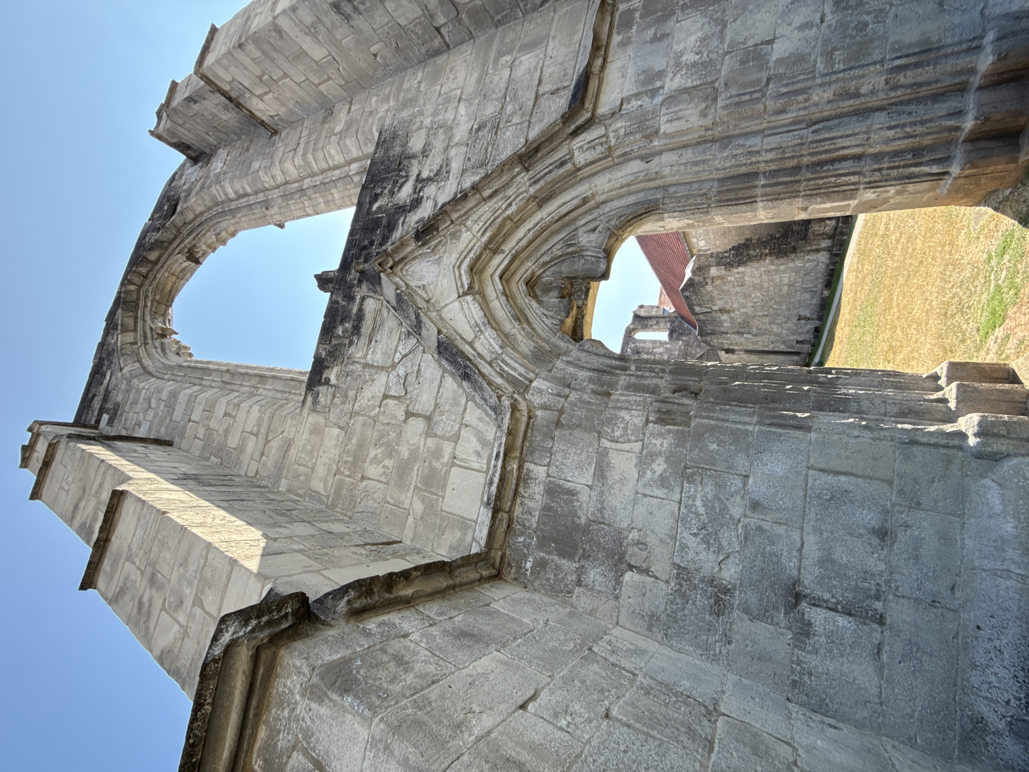 An upward-angle, full shot captures the stone ruins of a church in Walkenried, Germany, on a clear day. The sun illuminates the weathered stone walls, highlighting the architectural details of Gothic arches and large, empty window frames. A patch of dry, yellow grass and some green grass lie in the foreground, with a path leading through the ruins. In the background, a section of a building with a red tile roof is visible. The sky is a bright, clear blue.