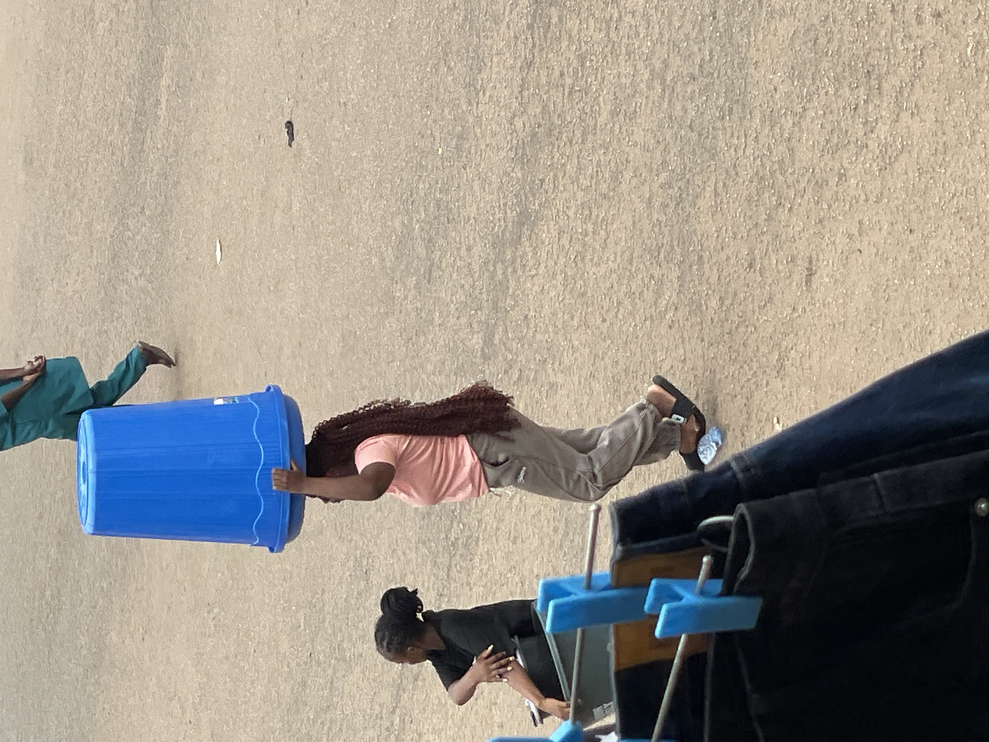 The image shows a daytime scene in Bukuru, Nigeria. A young woman in a pink shirt and grey pants is placing a large blue plastic trash can over her head. Another person, wearing green clothing, stands to the side of the can. A second woman, wearing a black shirt, is present near a clothes rack displaying jeans. The setting appears to be an outdoor area with a paved ground surface.