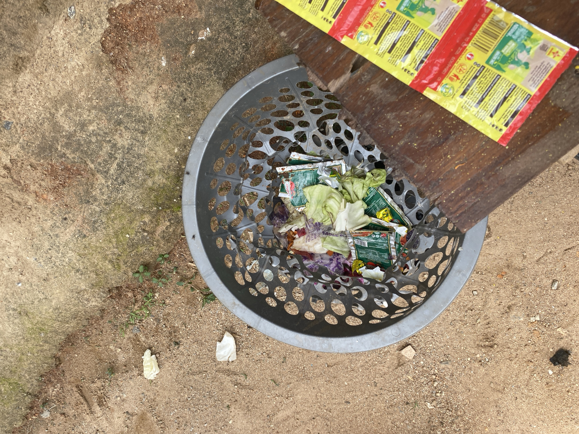 This image depicts an outdoor waste accumulation area in Kubwa, Nigeria, viewed from a high-angle perspective. A grey, perforated plastic laundry basket, serving as a trash receptacle, rests on a light brown, sandy ground intermixed with small pebbles. Inside the basket are various discarded items, including green and purple vegetable leaves, likely cabbage, and numerous small, crinkled plastic sachets, some with green and red designs.
To the left of the basket, on the sandy ground, are scattered small green plants and two small, irregular white pieces of material, possibly plastic or paper. A darker, moss-covered concrete surface occupies the upper right quadrant of the image. A dark wooden surface, possibly a plank or tabletop, extends from the right edge, partially overlapping the basket. On this wooden surface lies a rectangular yellow and red plastic package, featuring green graphic elements and a visible barcode. No individuals are present in the scene.