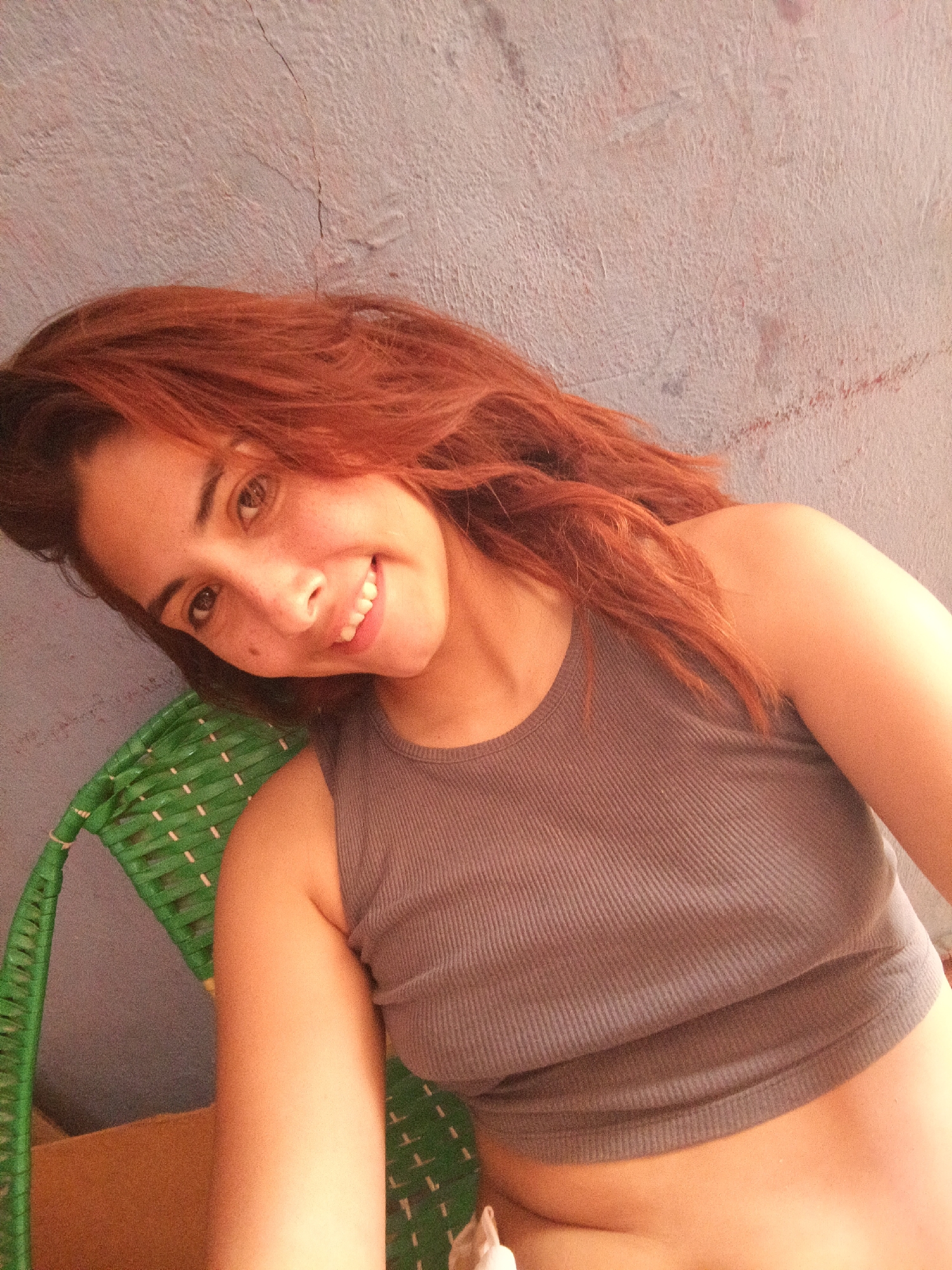 A woman with red hair is sitting in a green wicker chair. She is wearing a gray ribbed tank top. The background is a light purple wall with some cracks and discoloration. The location is Unknown, Unknown.