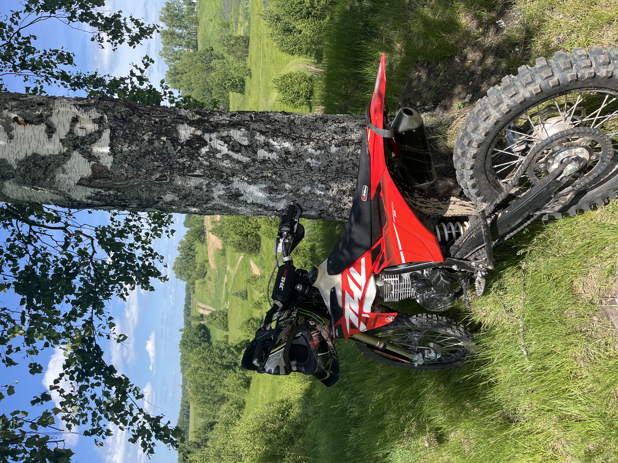A red and white off-road motorcycle is parked, leaning against the trunk of a large birch tree on a sunny day in Новочувашский, Russia. The motorcycle, identified by "JHLR" branding on its handlebars and a "JHC" logo on its tank shroud, displays mud on its knobby tires and frame, indicating recent use. A grey and black motocross helmet rests on the handlebars. The prominent birch tree, positioned centrally, exhibits characteristic white bark with dark fissures and shadowed areas. Its green leaves from overhead branches frame the upper view of a blue sky with scattered white cumulus clouds. The immediate foreground consists of tall, verdant green grass interspersed with areas of sun and shadow. The midground features rolling green hills covered in dense deciduous trees and open grassy patches, through which multiple dirt tracks or trails are visible winding into the distance. The far background presents a hazy outline of what appear to be industrial structures or towers on the left horizon, beyond more distant tree lines. No individuals are present in the scene.