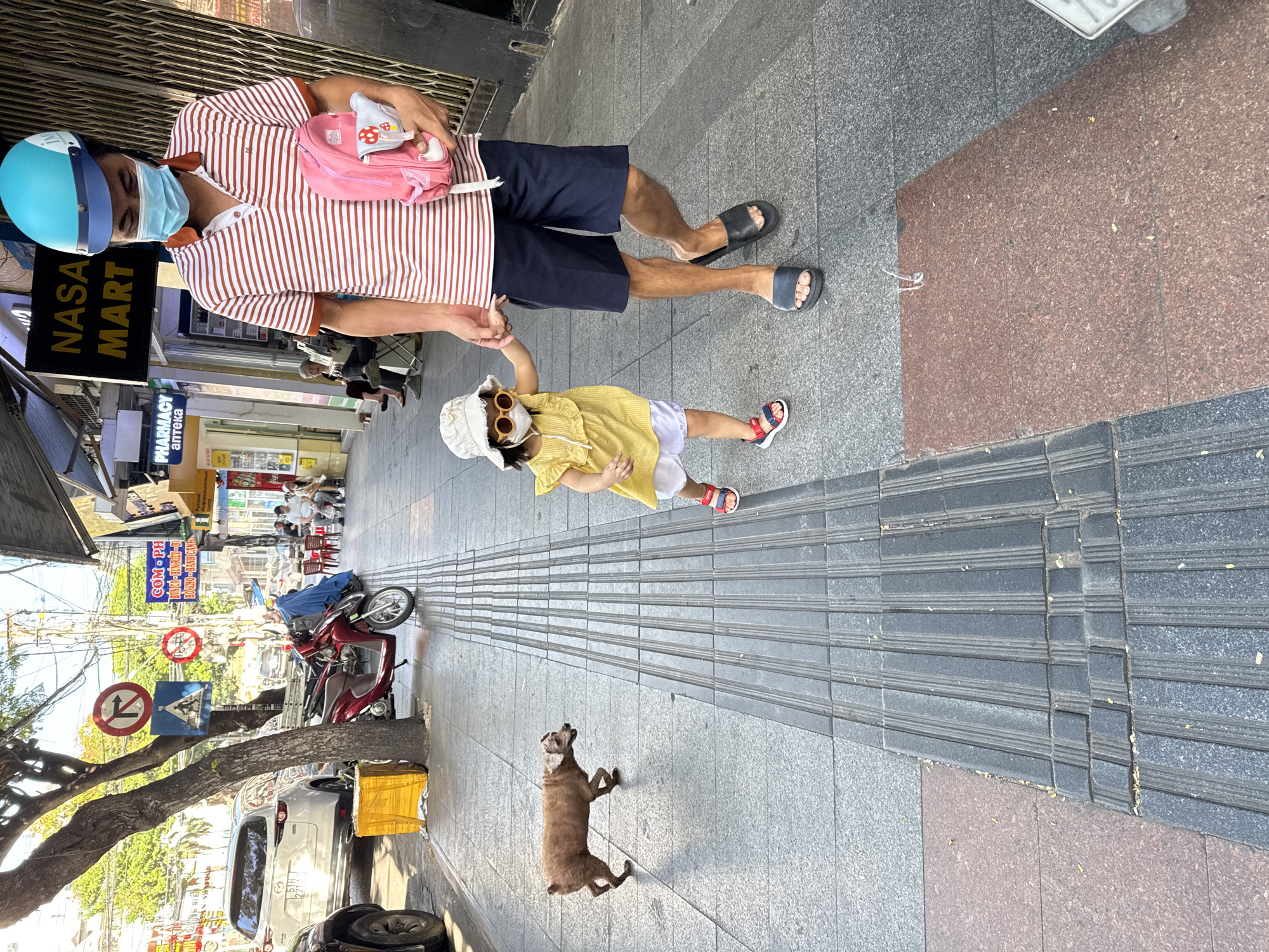 The image depicts a street scene in Nha Trang, Vietnam. A man wearing a striped polo shirt, dark shorts, a blue and cyan helmet, and a light blue face mask walks hand-in-hand with a young child. The child is wearing a yellow dress, white shorts, round sunglasses, and a white hat. The man carries a pink backpack. They are walking on a textured sidewalk. To their left, a small brown dog is walking away from the camera. In the background, there is a red scooter, a white SUV, and various shop signs, including one that reads "NASA MART." The environment appears to be a commercial street with buildings lining the sidewalk.