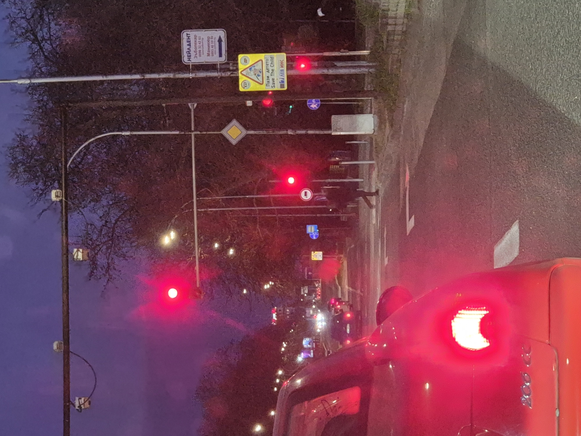 The image, likely taken in the city of Varna, Bulgaria, shows a red car stopped at a traffic light. The light is red, and there are multiple other traffic lights along the street, all also red. A pedestrian is standing near the crosswalk. Several road signs are visible, including one that reads "Пази детето! Save The Child!" ("Beware of the children" in Bulgarian), and a sign advertising a dentist and manicure services, "НЕЙЛДЕНТ". The environment is urban, with streetlights and trees lining the road. The sky is transitioning from dusk to night.