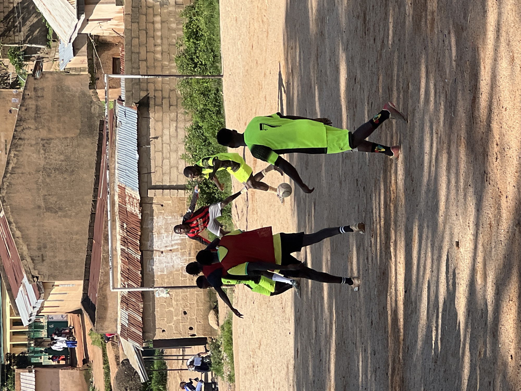 The image depicts a group of young men playing soccer on a dusty field in Jos, Nigeria. The scene is characterized by a rough, unpaved surface with visible shadows from nearby trees. In the background, there are several buildings with corrugated iron roofs, typical of many residential areas in Nigerian cities. A makeshift goalpost, constructed from metal pipes, stands at one end of the field.
Several players are actively engaged in the game. One player, wearing a bright green jersey with the number '7' on the back and black shorts, is prominently featured in the foreground. Another player in a red bib and black shorts is facing away from the camera. A third player, also in a green jersey, is in the middle ground, kicking a soccer ball. A fourth player, wearing a red and black striped jersey and white shorts, is also visible, possibly a goalkeeper. Other players are partially visible in the background.
The environment suggests a casual, community-based sporting event. The presence of simple, functional buildings and the unpaved field indicate a local setting, likely a neighborhood pitch. The overall atmosphere is one of active recreation, with the players focused on the game. The sunlight casts long shadows across the field, indicating it is likely late afternoon.