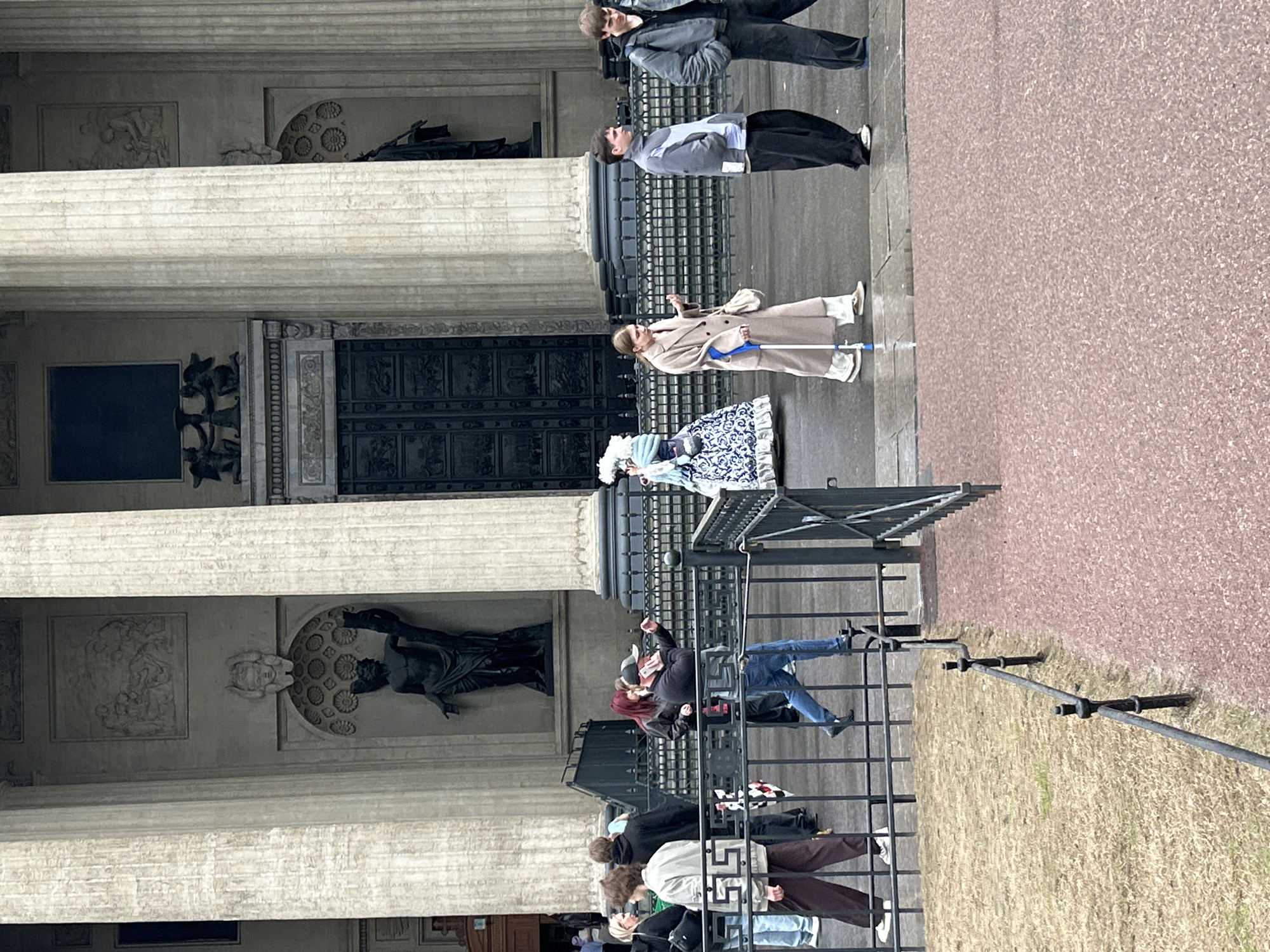The image captures a scene in Saint Petersburg, Russia, in front of a grand building with classical architecture. The building's façade features large columns and sculptural elements. People are present, including tourists and possibly costumed performers. Some individuals are walking, standing, or interacting. The environment includes the building's architectural details, the pedestrian area, and a glimpse of foliage.