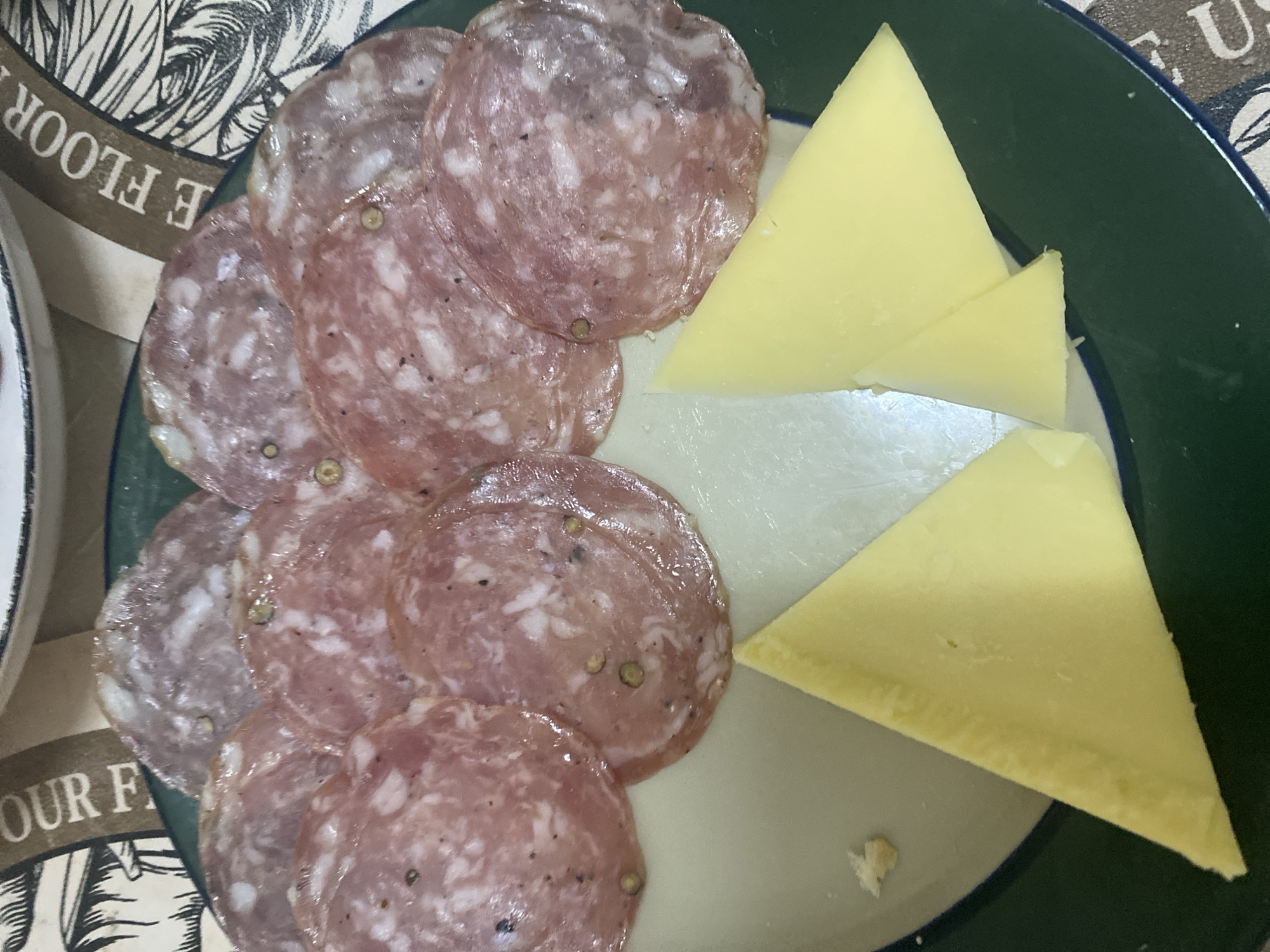 This close-up image presents a serving of charcuterie and cheese on a plate, set in Baan Chaloklum, Thailand. The main subjects are thinly sliced salami and triangular pieces of cheese.
On the left side of the plate, several round slices of salami are arranged in an overlapping pattern. The salami is pinkish-red with visible white marbling of fat, characteristic of cured meat. Numerous dark specks, likely black peppercorns, and some lighter, possibly green peppercorns are embedded within the slices.
To the right, three distinct triangular pieces of pale yellow cheese are arranged. The cheese appears firm and uniformly colored. Two larger triangles are placed towards the top and bottom right, with a slightly smaller one in between.
The food is presented on a round plate with a dark green rim and a lighter, possibly off-white or light grey, central eating surface. A portion of another white plate or bowl is partially visible on the far left edge of the frame.
The background features a patterned surface, which appears to be a placemat or tablecloth. Visible text on this surface includes the fragmented words "E FLOOR" and "OUR F", presented in a decorative font, alongside graphic elements that resemble foliage or a stylized design. The lighting is bright and even, suggesting an indoor setting or a well-lit area. There are no people visible in the image. The arrangement suggests a simple appetizer or snack.