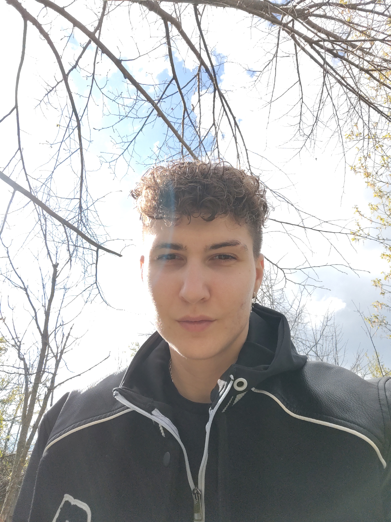 A selfie of a person with short curly brown hair, wearing a black jacket with white trim, is taken outdoors with bare tree branches framing the top of the image against a sky with scattered clouds. The location is Unknown, Unknown.