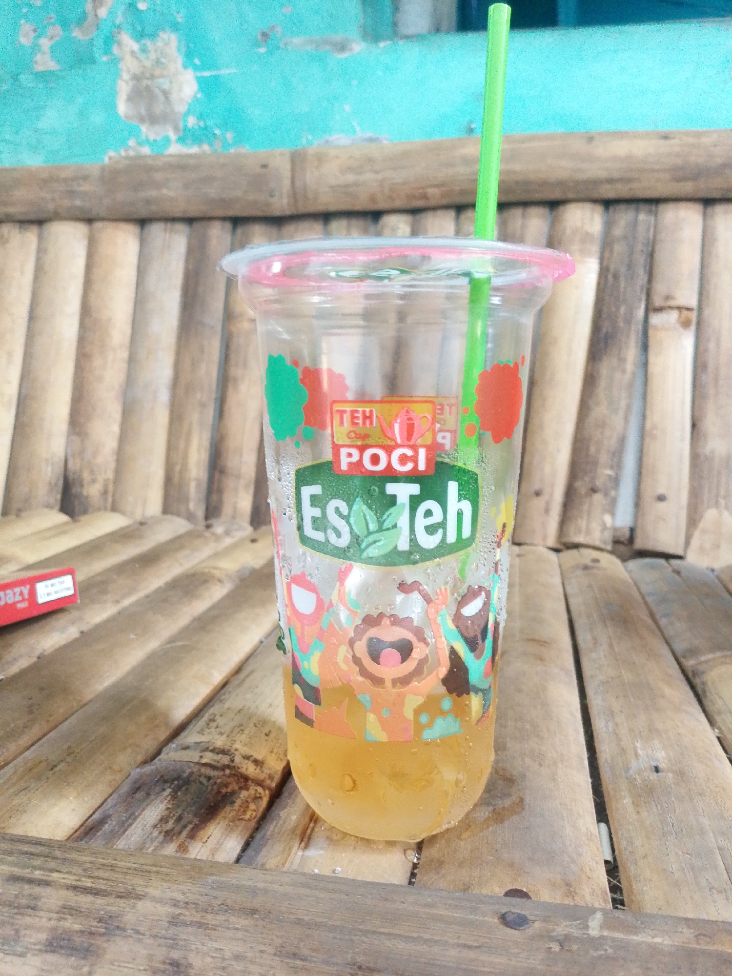 A plastic cup of Teh Poci iced tea sits on a bamboo bench in Tasikmalaya, Indonesia. The cup is clear with a pink lid and a green straw. The label on the cup features cartoon characters and the words "TEH POT", "POCI", and "Es Teh". The background is a teal-colored wall with peeling paint, and the bench is made of weathered bamboo slats. A red and white cigarette pack is partially visible on the left side of the image. The scene appears to be outdoors, possibly in a cafe or roadside stall, given the casual seating and rustic elements. The lighting suggests it is daytime.