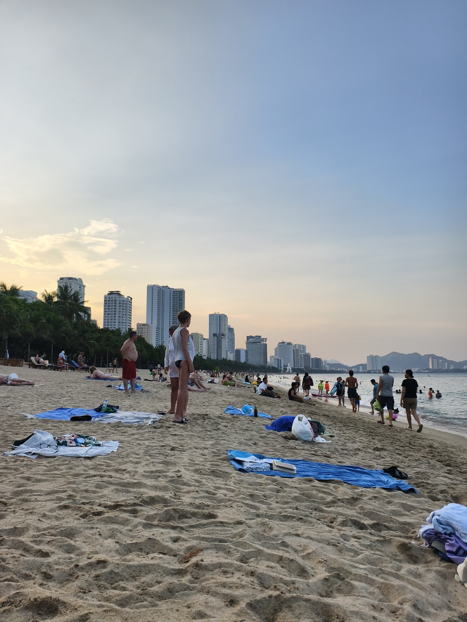 The image shows a crowded beach in an urban area. People are engaged in various activities such as swimming, walking along the shore, sunbathing, and standing around. Beach towels and personal belongings are scattered on the sand. In the background, there are numerous high-rise buildings. The sky is partly cloudy with a hint of sunset colors. The location appears to be a coastal city, with mountains visible in the distance.