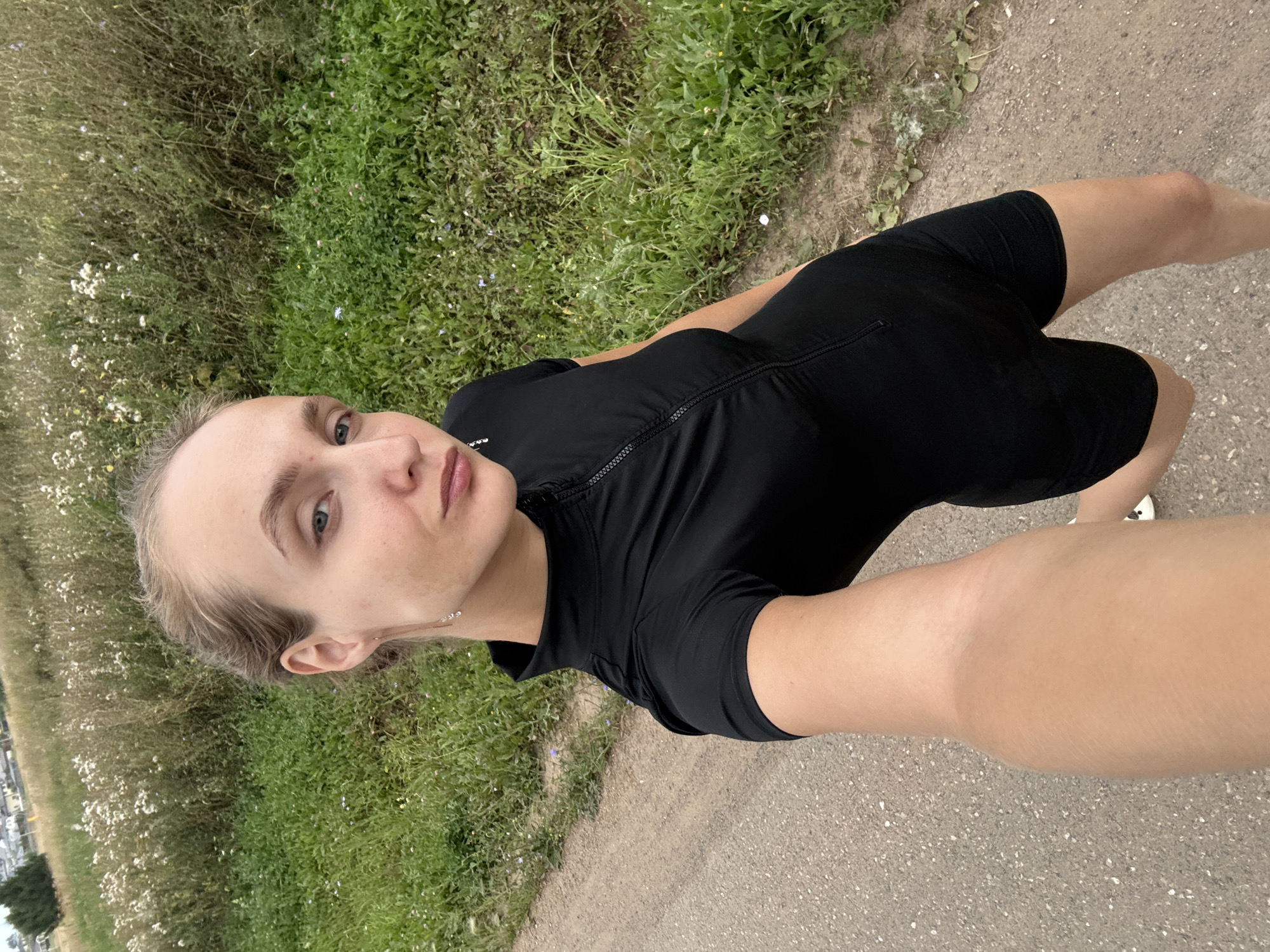 A blonde woman is taking a selfie outdoors. She is wearing a black, short-sleeved, zippered top and black shorts. She has on earrings and her hair is pulled back. She is standing on a gravel path next to a grassy field with wildflowers. The background shows more grassy fields and some distant buildings. The approximate location is Podshivalovo, Russia.