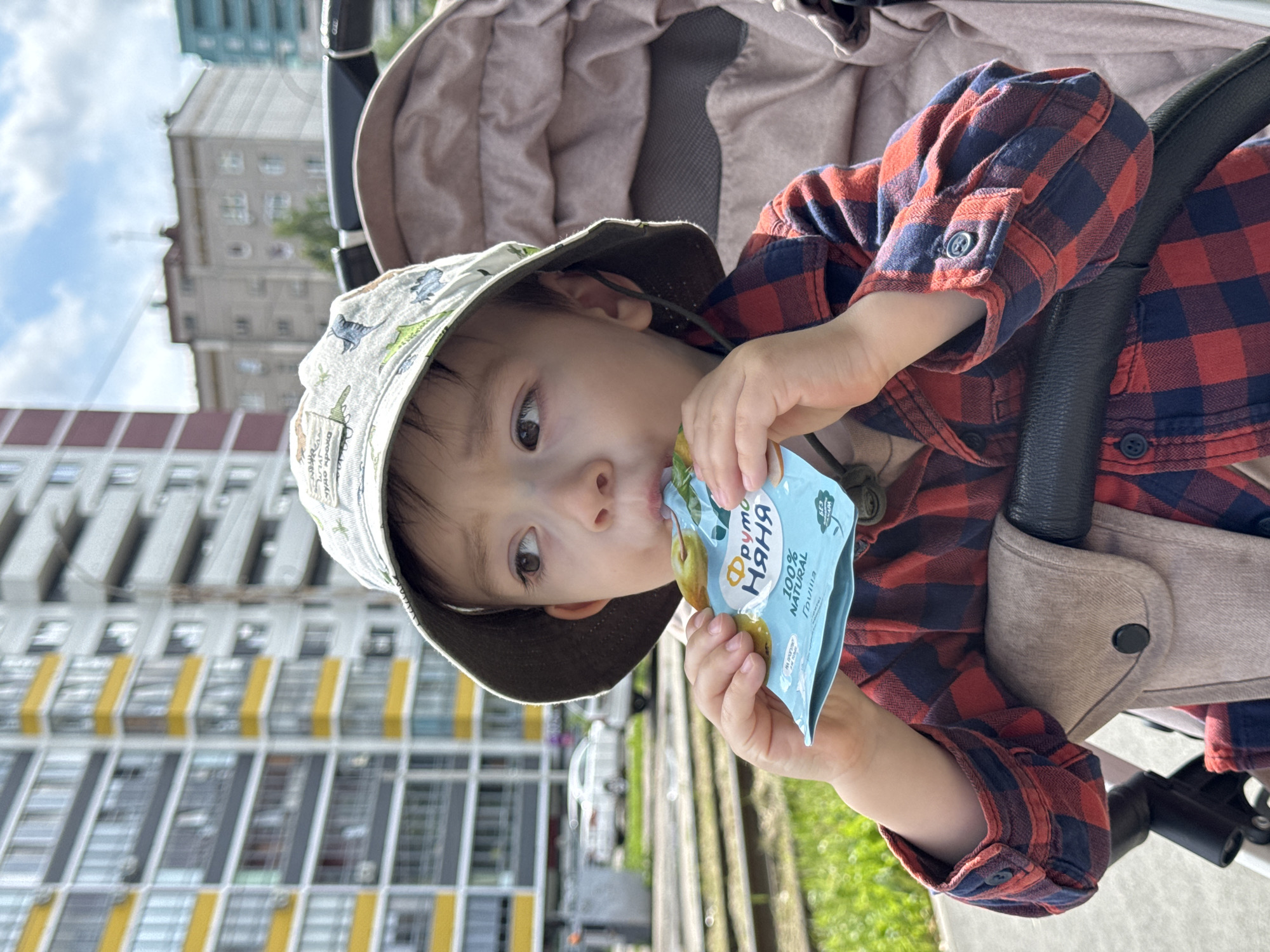 A young child wearing a bucket hat and a red and blue checkered shirt sits in a stroller and eats from a pouch. The child's face is turned to the right, and their eyes are focused on the pouch. The stroller is a light brown color, and the child's arms are resting on its frame. In the background, there are tall buildings and a clear blue sky with some clouds. A white car is parked on the street in front of the buildings. The location is the city of Izhevsk, Russia.
