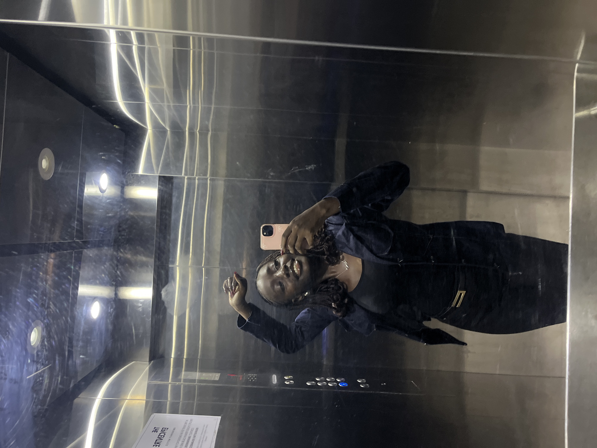 A woman is taking a selfie in an elevator. She is holding a pink cell phone. She is wearing a dark-colored outfit, possibly a jacket and top. The elevator has stainless steel walls and a control panel. The environment suggests a modern building in Karum, Nigeria. There is a sign present in the elevator, possibly indicating building information.