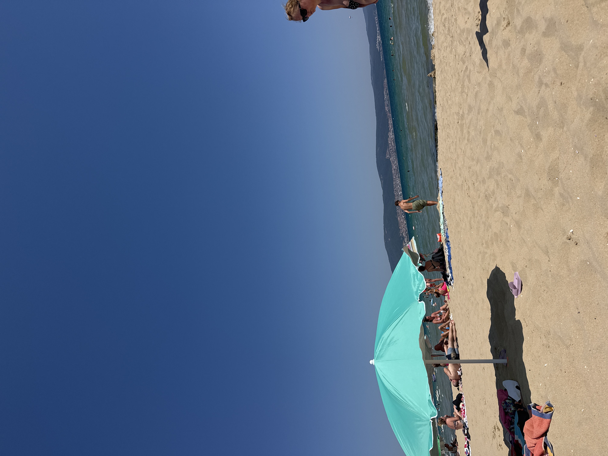 The image depicts a sunny day at a beach in Nesebar, Bulgaria. The foreground is dominated by golden sand with several beach towels and flip-flops scattered. A large, turquoise beach umbrella provides shade for a group of people, some lying down and others sitting. In the background, the Black Sea is visible with calm waters, and a distant coastline with buildings and hills can be seen under a clear blue sky. Several people are in the water, and a young man in shorts stands near the water's edge. On the right side of the frame, a woman wearing sunglasses and a bikini is looking away from the camera. The overall scene conveys a relaxed atmosphere of a typical summer day at the beach.