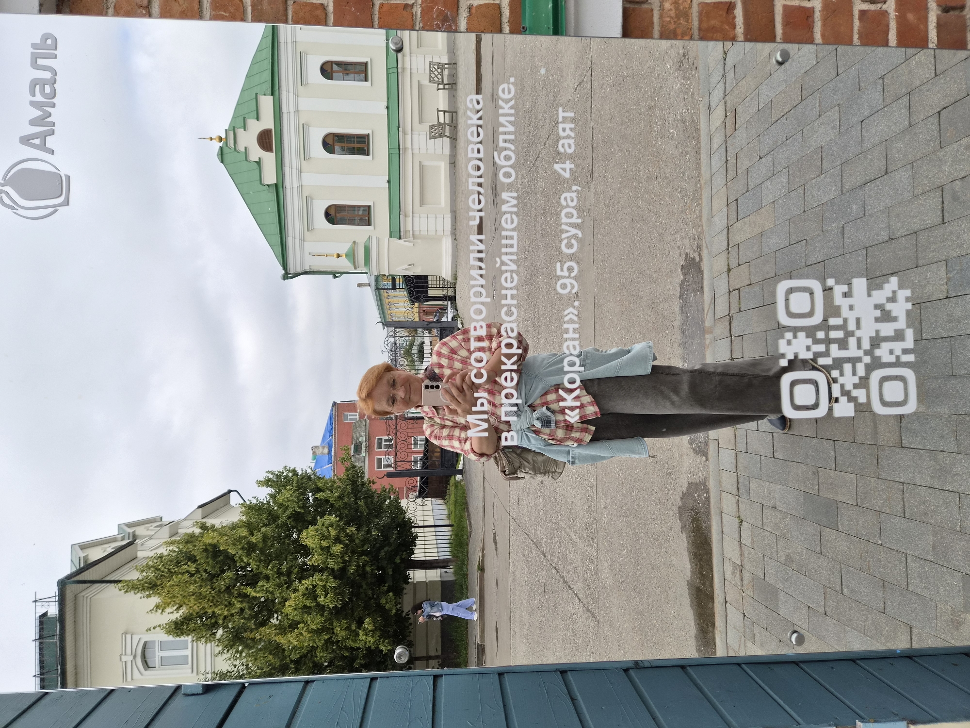 A woman with reddish-blonde hair, wearing glasses, a red and white checkered shirt, and dark pants, stands in front of a mirrored surface, holding a smartphone to take a selfie. The mirror reflects the woman, a QR code, and text in Russian. The text reads: "Мы сотворили человека в прекраснейшем облике. «Коран», 95 сура, 4 аят" which translates to "We have created man in the finest form. 'The Quran', Chapter 95, Verse 4".
The reflection also shows a building with a green roof and white walls, several trees, and a cloudy sky. A person in a denim jacket and light-colored pants is visible in the background walking away from the mirror.
The scene appears to be in an outdoor urban setting, likely in Kazan, Russia, given the Cyrillic text and architectural style. The mirrored surface is likely part of a shop or exhibition display, as indicated by the brand logo "Амаль" (Amal) with a stylized flask symbol at the top of the mirror. The ground is paved with grey interlocking bricks. The blue wooden structure on the left side of the frame suggests the presence of a building or a stall.