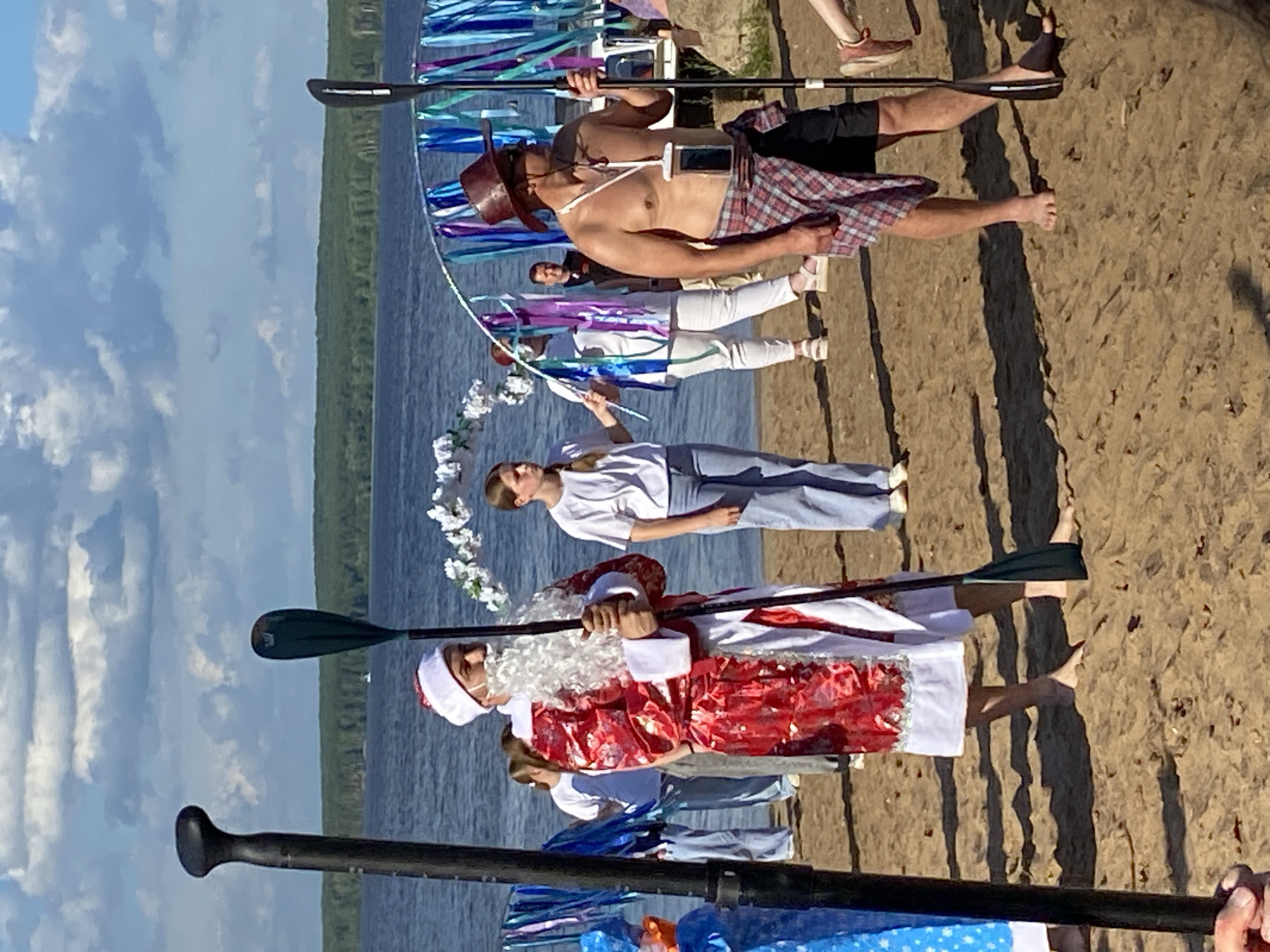 On a sandy beach next to the Kama River in Chaikovsky, Russia, people are gathered. A man dressed as Santa Claus, complete with a white beard and red suit, stands holding a paddle. Nearby, a shirtless man in a hat and shorts, with a plaid shirt tied around his waist, also holds a paddle. Other individuals in casual clothing are visible in the background, some near a decorative archway adorned with flowers and ribbons. The sky is partly cloudy, and a forest line is visible across the river.