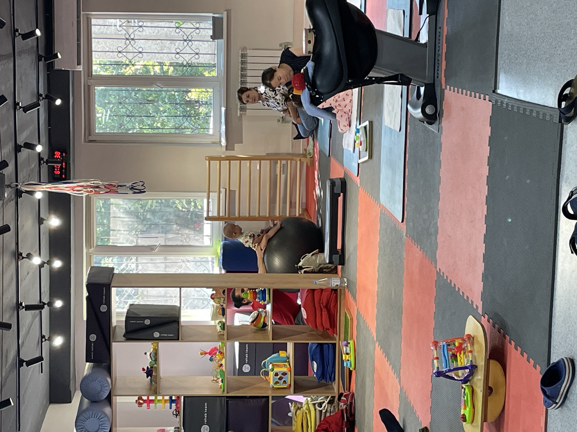 The image depicts the interior of a rehabilitation center in Almaty, Kazakhstan. The room is furnished with various therapeutic and recreational equipment. In the foreground, a black exercise ball is placed on a gray mat, with a baby sitting on top of it. To the right, two women are seated on yoga mats, engaging in an activity. One woman is wearing a black and white patterned shirt, while the other wears a dark blue shirt.
The environment includes shelves filled with children's toys and rehabilitation aids. A wooden climbing frame is visible against the wall, and a digital timer displays "8:08:38" on the ceiling. Windows with decorative metal grates allow natural light into the room. The floor is covered with interlocking foam mats in red and gray. A specialized exercise machine with a black seat is positioned in the right-hand side of the frame. Multiple pairs of footwear are scattered on the floor. The overall scene suggests a therapeutic session or class for children and adults.