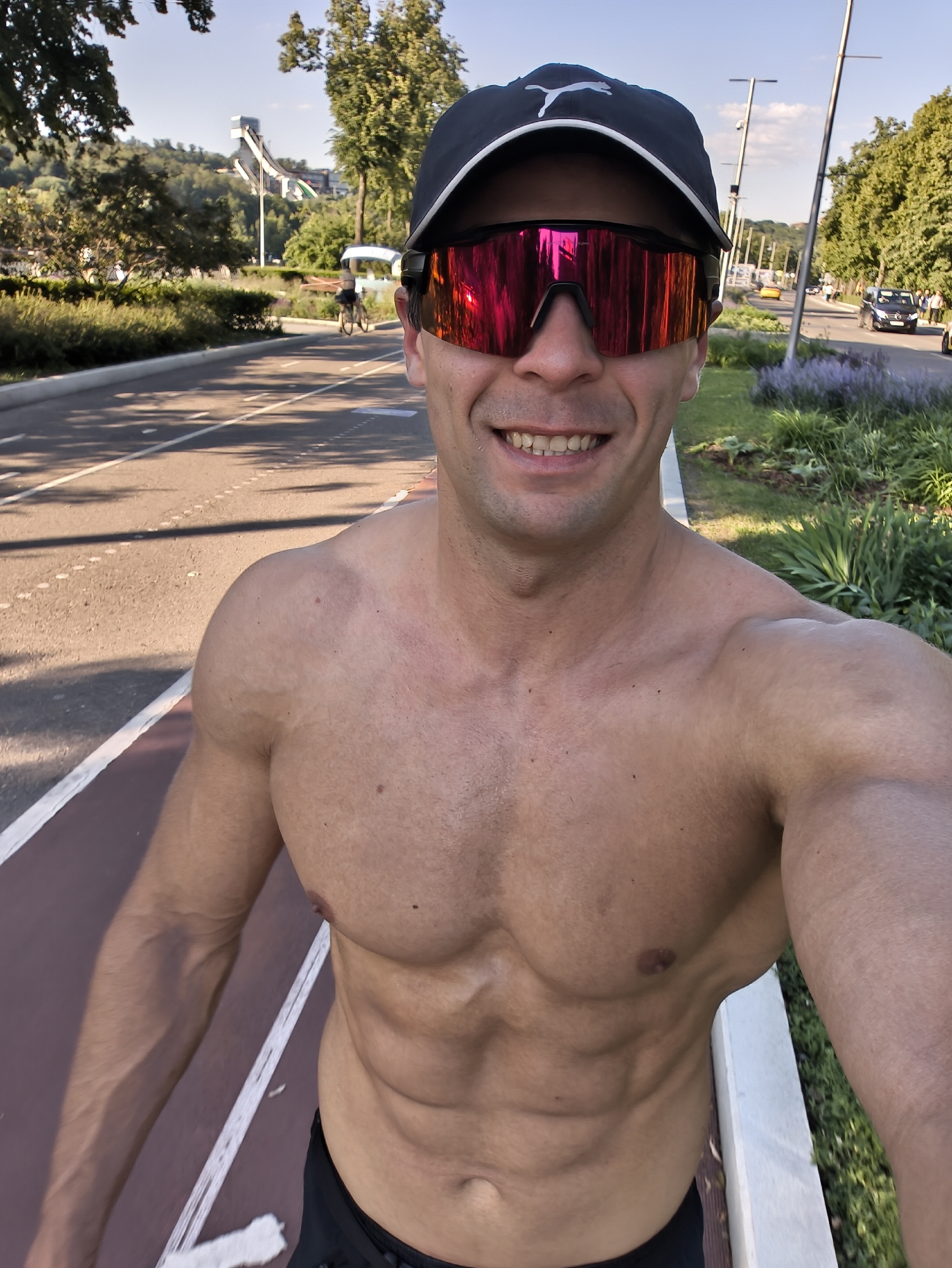 This is a selfie taken by a shirtless man wearing a black baseball cap and reflective red sunglasses. He has well-defined abdominal muscles and is smiling. He is standing on a red paved path, possibly a running or cycling track, with white lane markers. Behind him, a road with traffic can be seen, including a black van and a yellow car. Trees and a green grassy area with purple flowers line the road. In the background, a ski jump structure is visible on a hill. The sky is blue with some clouds, suggesting it is daytime, likely late afternoon due to the shadows. The scene appears to be outdoors in a park-like setting within a city. The man's posture and expression suggest he is active and enjoying himself. The location context provided is Moscow, Russia. There is a visible logo on the cap that appears to be a white Puma cat logo.