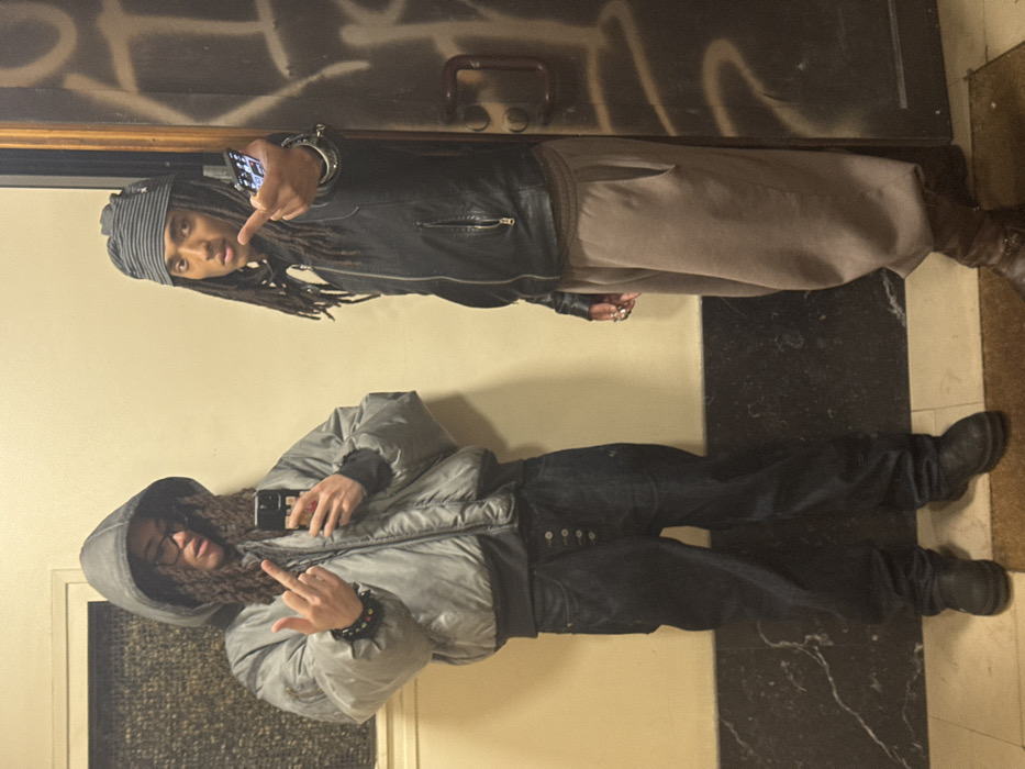 Two people pose for a photo. The individual at the top, with dreadlocks and a striped head covering, wears a black leather jacket and tan pants. They hold a phone and make a hand gesture. The person below wears a hooded, light grey puffer jacket and dark jeans, also holding a phone while displaying a different hand gesture. Both wear dark shoes. The backdrop features a wall with possible graffiti and a black and white patterned floor.